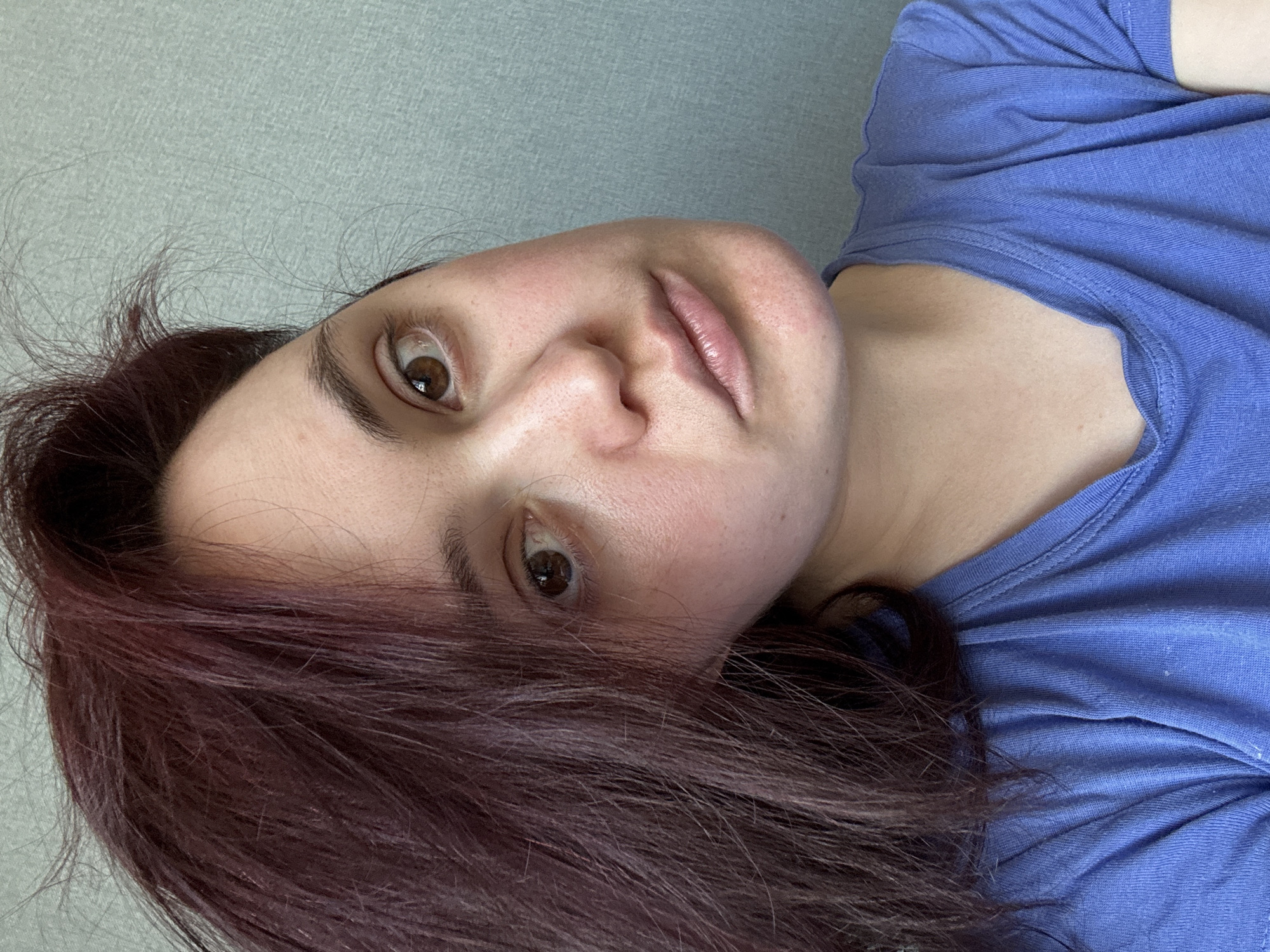 A woman is depicted in a close-up, angled selfie. She has fair skin, brown eyes, and dark auburn hair. She is wearing a blue t-shirt. The background is a plain, light grey wall. There is no evidence to suggest the location of the photo is in Izhevsk, Russia.