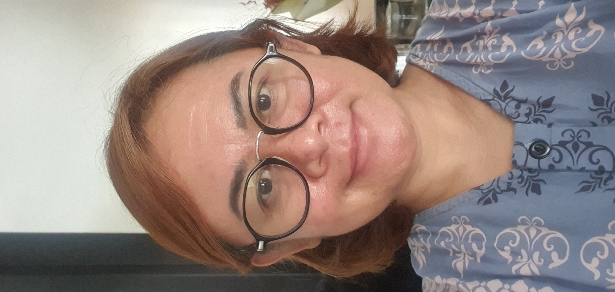 A medium shot selfie features a woman with shoulder-length auburn hair and fair skin. She is wearing round, dark-rimmed glasses and a blue and white patterned shirt. Her expression is a slight smile. The background is out of focus, showing a hint of a white wall and dark vertical structure on the left. The image appears to be taken indoors.
