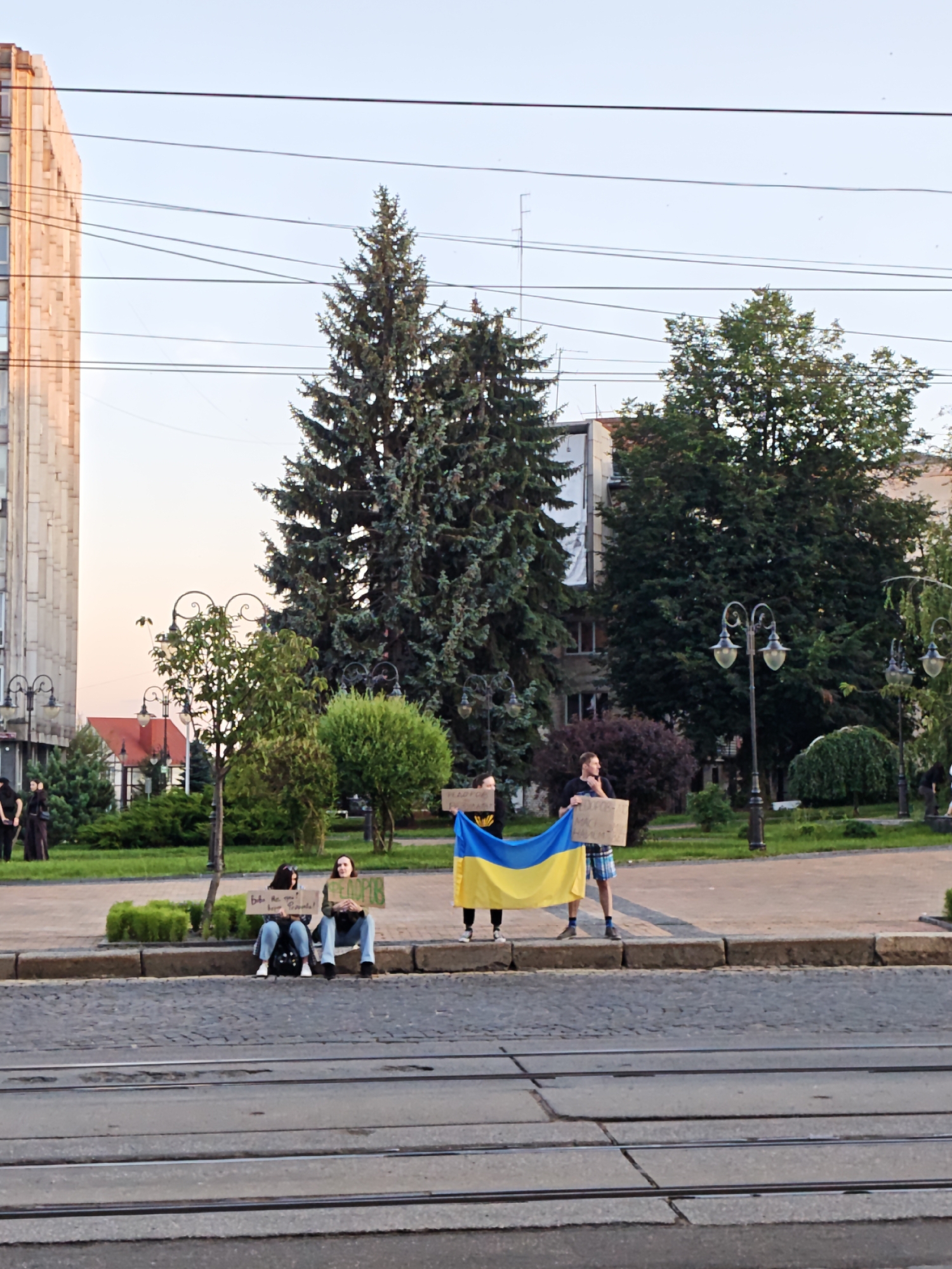 The image depicts a group of people holding Ukrainian flags and signs in an urban setting. The background features a building with light-colored facade and numerous trees, including several large evergreen trees. The foreground shows a street with tram tracks. Several individuals are visible, some holding signs and a large Ukrainian flag. Two women are seated on a curb, each holding a sign. One sign appears to have Ukrainian text. A man stands holding a sign and a portion of the Ukrainian flag. The sky is clear and the lighting suggests it is either early morning or late afternoon. The presence of tram tracks and the architectural style of the building suggest an urban environment in Ukraine. Without additional context, the specific location in Vinnytsia cannot be precisely identified, but the scene is characteristic of a public space within the city.