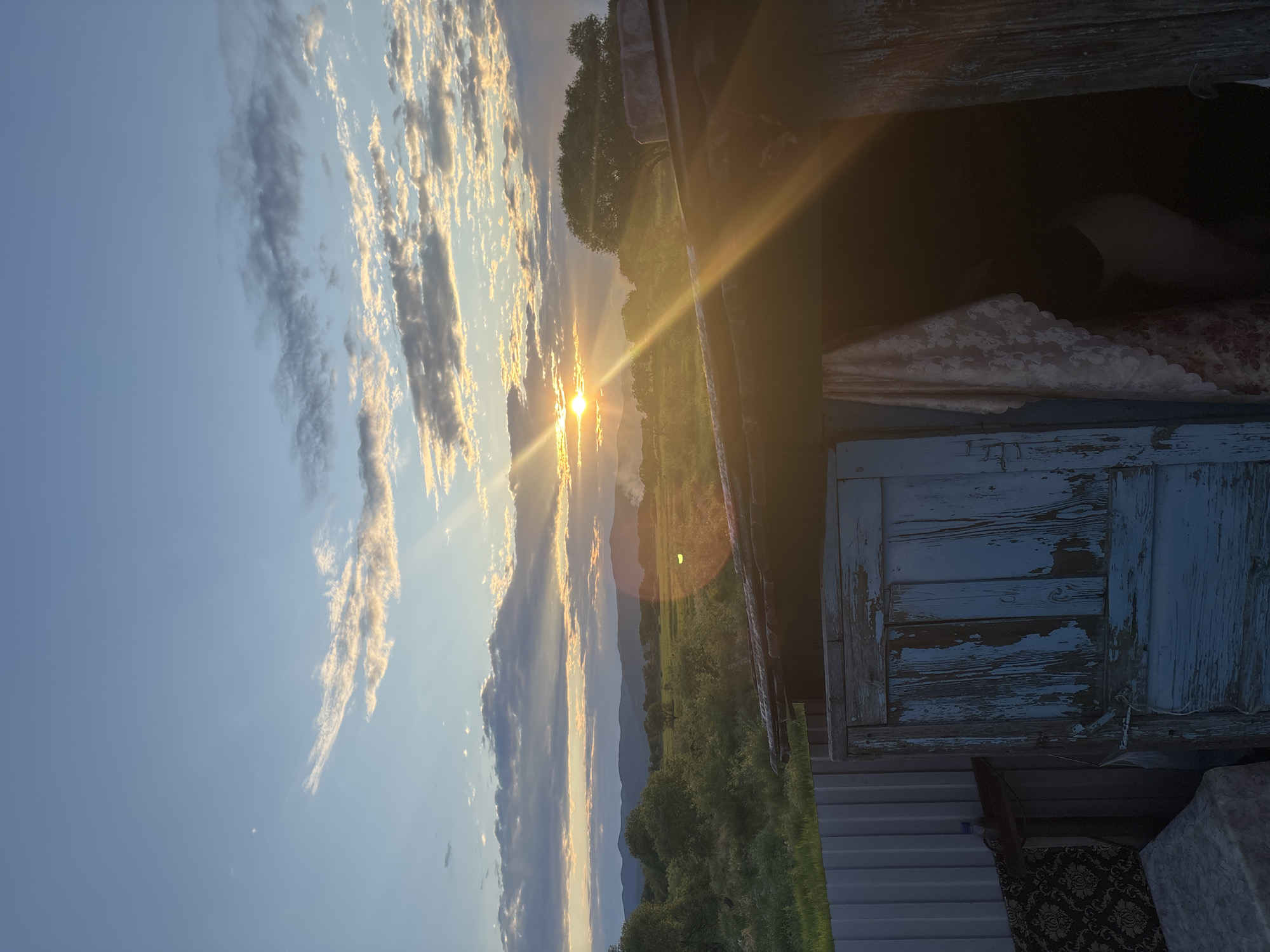 A bright sun sets behind rolling hills and a vast, green field. The sky is a brilliant blue, streaked with wispy white and grey clouds. Beams of golden sunlight pierce through the clouds, creating a radiant halo effect around the sun. In the foreground, the dilapidated roof of a rustic wooden structure partially obscures the view, with a weathered blue door in its opening. A pale, patterned fabric hangs inside the structure. A corrugated metal fence runs along the left side of the frame, separating the foreground from the field. The overall scene depicts a serene, rural sunset.