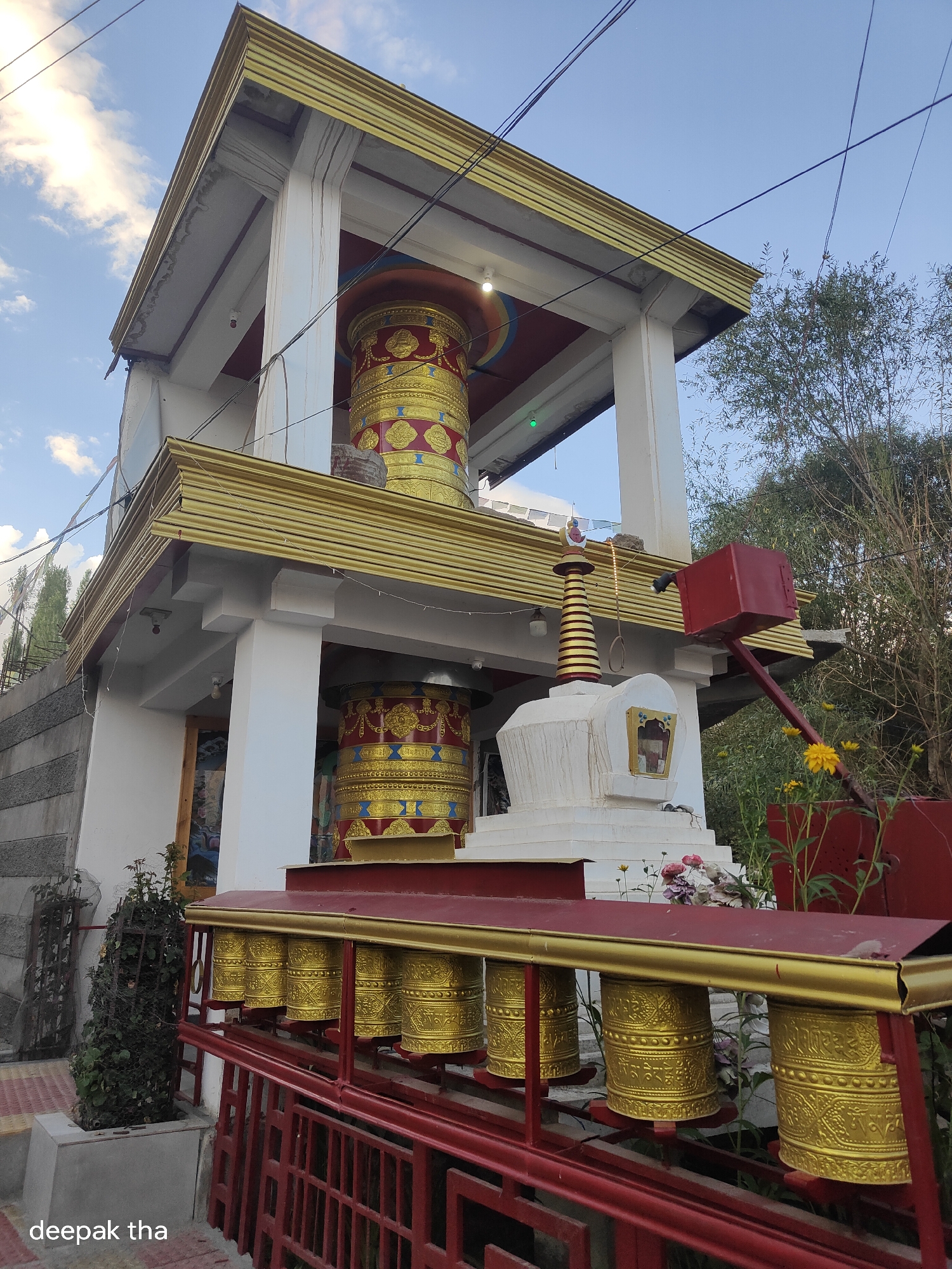 This image depicts a Buddhist stupa and prayer wheels located in Leh, India. The stupa is a white, tiered structure with a golden spire. Several large, cylindrical prayer wheels, adorned with intricate golden designs and inscriptions, are arranged in a row in front of the stupa, mounted on a red and gold railing. Behind the stupa, a multi-story building with white pillars and decorative golden accents is visible. A red, hammer-like object is positioned on an extended arm to the right of the stupa. Yellow flowers and green foliage surround the religious artifacts and building. The sky above is blue with scattered white clouds. The image is a medium shot taken from a low angle, emphasizing the scale and grandeur of the Buddhist structures. A watermark with the text "deepak tha" is present in the bottom left corner of the image.