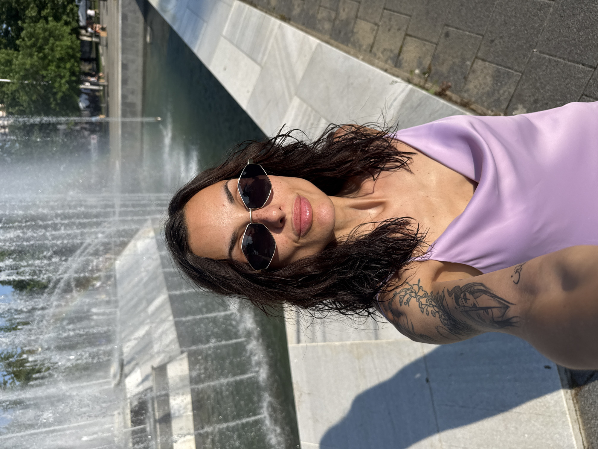The image displays a self-portrait of a woman positioned in front of The Singing Fountains in the City Garden of Pleven, Bulgaria, under direct sunlight. The subject has dark, damp-appearing wavy hair and wears dark hexagonal sunglasses with light frames. She is dressed in a light lavender satin cowl-neck camisole. A detailed black ink tattoo featuring a stylized face within a geometric design and a vine pattern is visible on her upper left arm. Her lips are slightly pursed. In the background, the large, multi-tiered fountain emits numerous jets of water, forming a dense spray. Beyond the fountain, green trees and indistinct figures of people are visible in the distance, characteristic of a public park environment. A light-colored stone curb separates the fountain from a grey paved walkway in the foreground. Strong shadows are cast on the ground due to the bright sun.