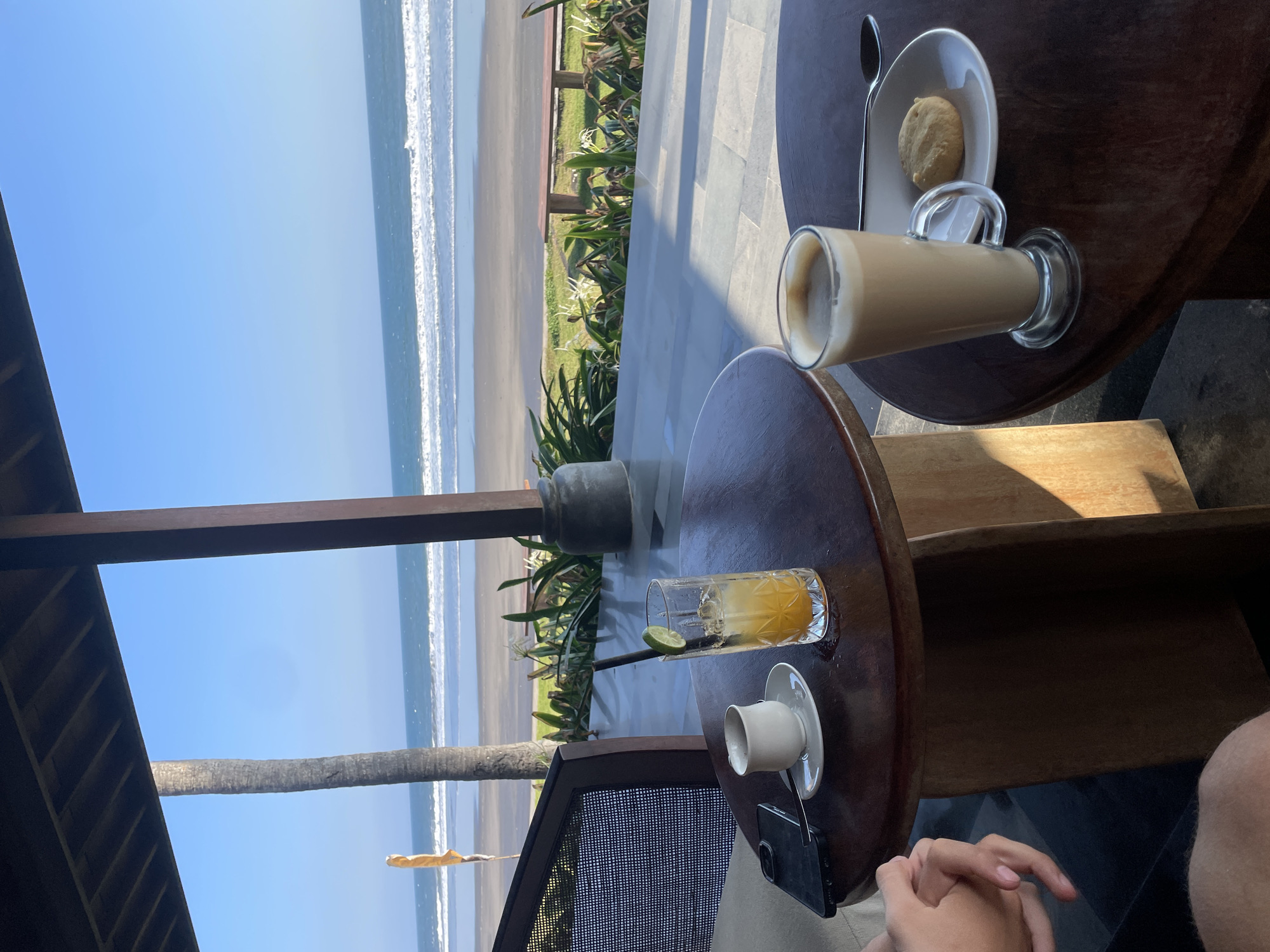 The image depicts a serene beachside cafe in Selemadeg Timur, Indonesia. Two wooden tables are set with drinks and a cookie. On the left table, a small white cup and saucer sit next to a tall glass filled with an orange beverage, garnished with a lime wedge and a black straw. A dark-colored smartphone is placed beside the cup. On the right table, a tall glass of coffee with a foamy top is presented with a single cookie on a white plate, accompanied by a spoon. In the background, the turquoise ocean meets a sandy beach, with gentle waves rolling towards the shore. Lush green foliage lines the edge of the beach, and a single palm tree stands tall against the clear blue sky. A covered patio area with wooden posts shades the cafe seating. A person's hands are visible in the lower left corner of the image, suggesting they are seated at the cafe.