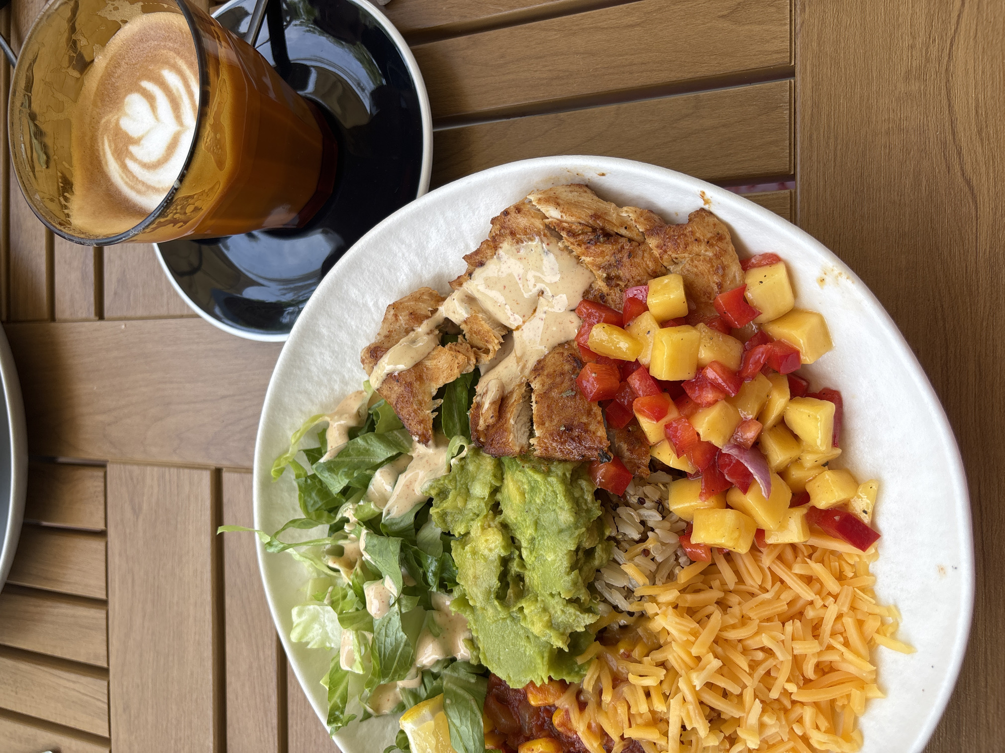 An overhead shot shows a white plate with a composed dish beside a coffee cup, sitting on a brown, slatted wooden surface. The dish on the plate is divided into sections: grilled chicken drizzled with sauce, a mango and red pepper salsa, shredded cheddar cheese, mixed grains, a scoop of guacamole, and salad greens with dressing. Beside the plate is a clear glass cup of coffee with latte art on a black saucer. The scene may be located in Choeng Thale, Thailand.