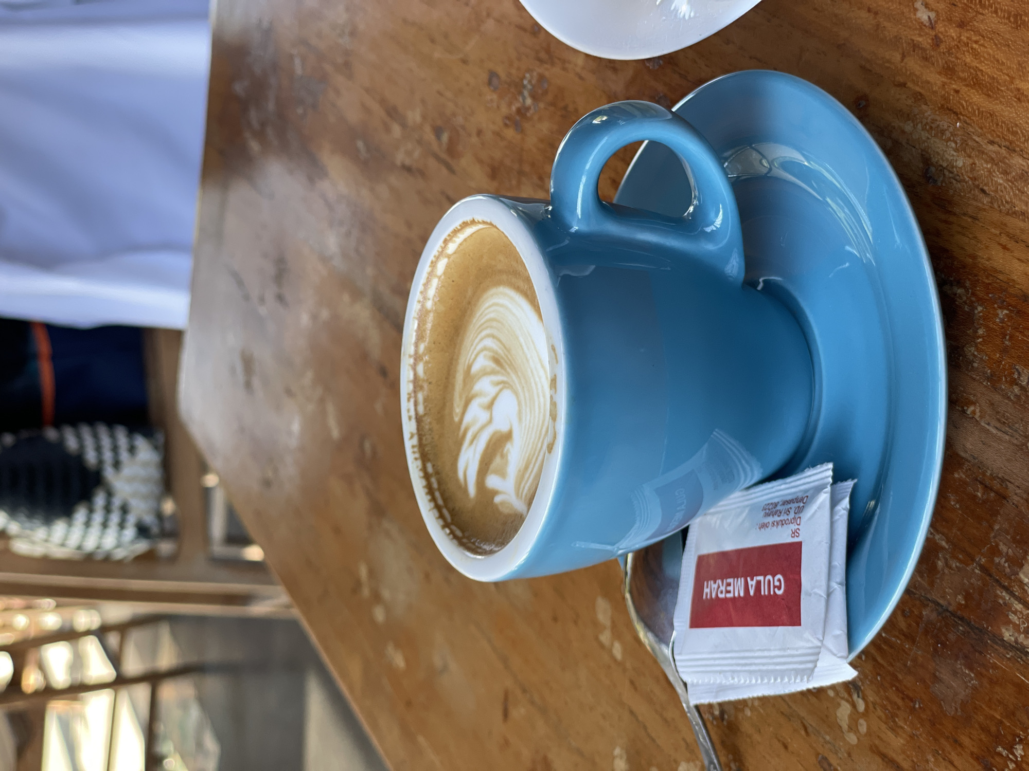 A close-up shot focuses on a blue ceramic cup of coffee with latte art, placed on a matching saucer atop a wooden table. A silver spoon rests on the saucer, alongside a packet of white sugar with red lettering. In the blurred background, elements of a cafe environment are visible, including chairs, a woven basket, and white fabric. The setting appears to be a cafe in Denpasar, Indonesia.