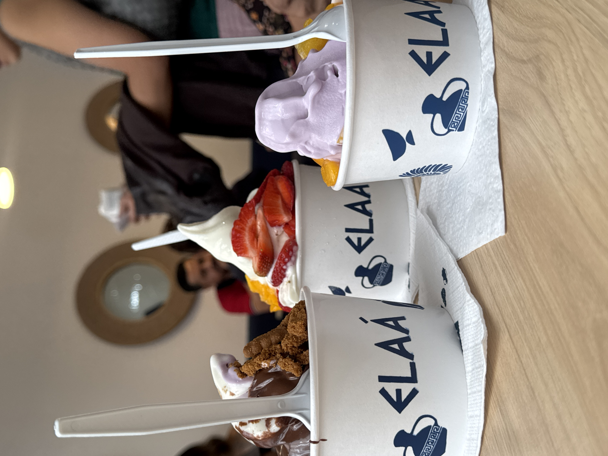 Three cups of frozen yogurt stand on a white napkin on a light wood-grain table, oriented diagonally. The cups are white with the name "ELAÁ" printed in blue. Each cup has a plastic spoon. The top cup contains purple frozen yogurt with yellow fruit pieces. The middle cup contains white frozen yogurt with sliced strawberries and yellow fruit. The bottom cup contains vanilla frozen yogurt with chocolate sauce and brown, crumbled toppings. Partial views of people are visible in the background, including one with a circular wall mirror behind their head. The location is Puebla, Mexico.