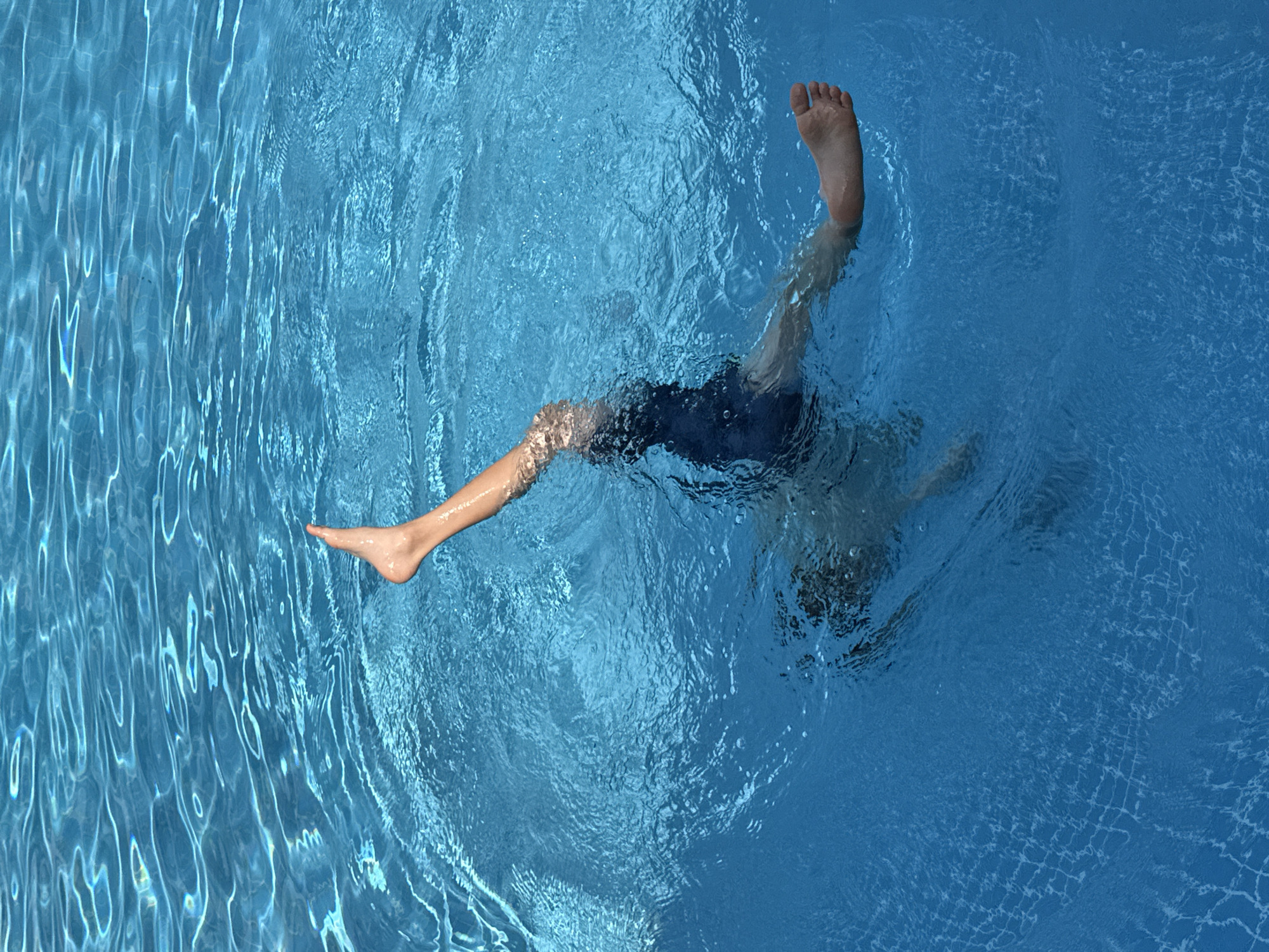 A person is submerged in a bright blue swimming pool, with only their legs and feet visible above the water. The person is in a vertical position, with their body upside down and legs extended upwards. One leg is bent at the knee, with the foot pointing towards the surface, while the other leg is more straightened, with the foot also visible. The water is clear and reflects the sunlight, creating ripples and patterns on the surface. The bottom of the pool is not visible. The scene appears to be in an outdoor swimming pool. There is no identifiable location within Dubai, United Arab Emirates, visible in the image.
