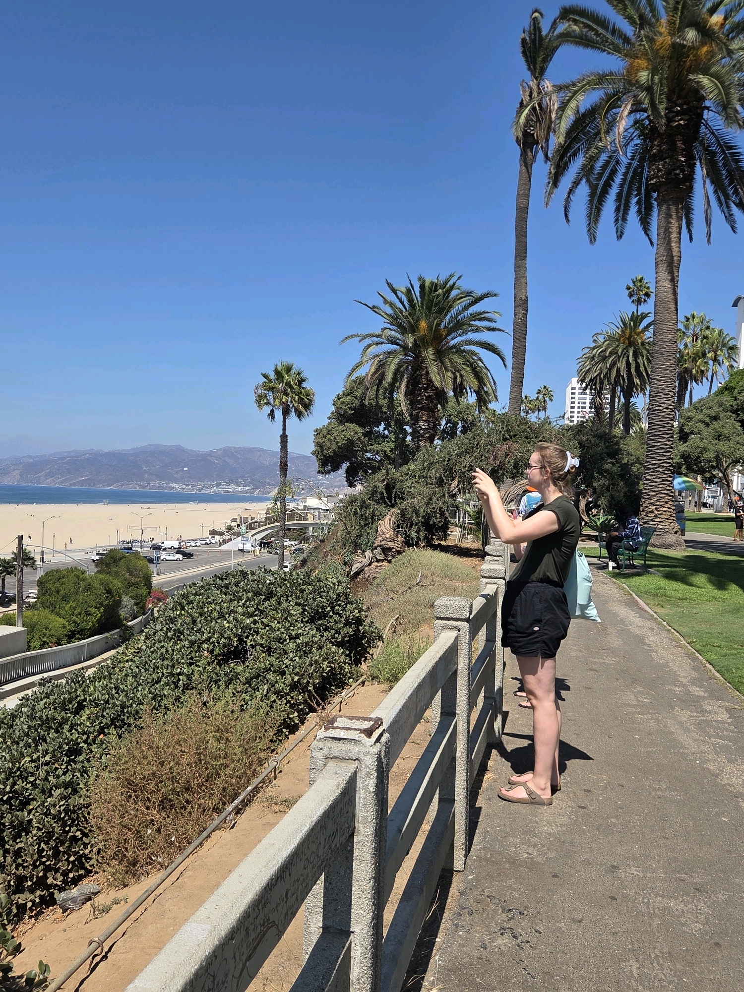 The image captures a sunny day at a coastal park in Santa Monica, California, likely near Palisades Park, offering a panoramic view of the Pacific Ocean and beach. In the foreground, a young woman with blonde hair tied up, wearing a dark green t-shirt and black shorts, stands on a paved walkway, holding a phone to take a picture. She is facing away from the camera, looking towards the ocean. A light blue bag is behind her, and her feet are clad in Birkenstock-style sandals. The walkway is bordered by a concrete railing with graffiti. Beyond the walkway, a lush green slope descends, populated with trees and bushes. Further in the distance, the wide sandy beach of Santa Monica is visible, dotted with small figures of people. Across the beach, a road runs parallel, with cars parked and driving. In the far distance, the vast blue Pacific Ocean stretches to the horizon, with a hazy mountain range forming the backdrop. Several palm trees are scattered throughout the scene, characteristic of the Southern California landscape. In the background, a tall, white building stands out amongst other structures along the coast, and a person is seated on a green park bench. The sky is clear and a vibrant blue.