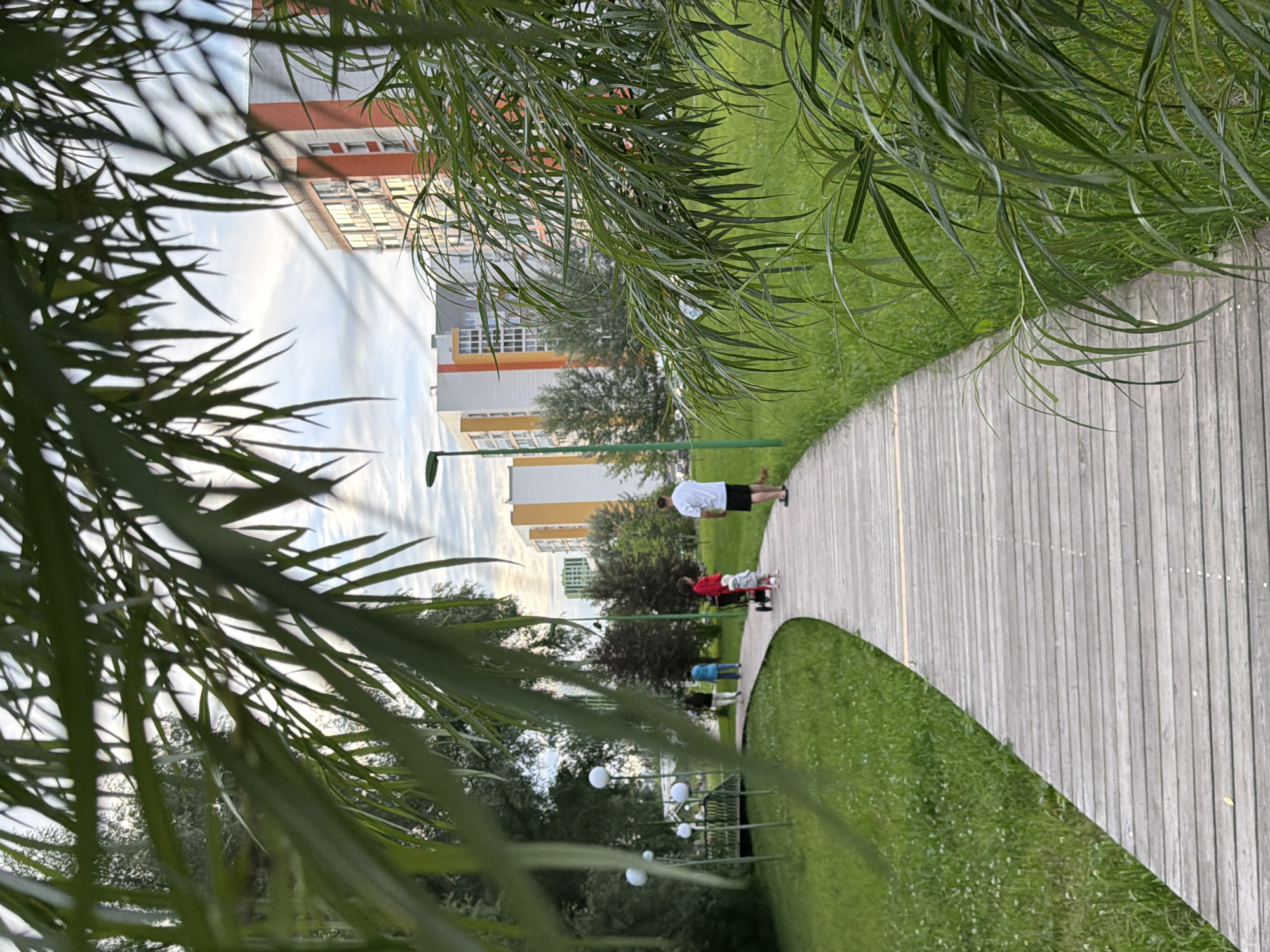 This is a vertical, medium-shot photograph taken in Kazan, Russia, depicting an outdoor scene with people on a wooden boardwalk. The image is partially obscured by the foliage of a tree in the foreground, with long, thin green leaves hanging down, creating a natural framing effect.
In the background, several multi-story buildings with light-colored facades and orange accents are visible. The sky above is partly cloudy. The boardwalk curves gently to the right, bordered by a well-maintained green lawn.
On the boardwalk, there are at least four individuals. The person closest to the foreground is a man wearing a white t-shirt and black shorts, facing away from the camera. Further along the boardwalk, a person in a red shirt is pushing a red wagon, with a smaller figure, possibly a child, in the wagon. Another person in a blue top is walking ahead. The people appear to be casually strolling or enjoying an outdoor activity.
Several white, spherical lights on green poles are positioned along the left side of the boardwalk, suggesting this is a park or recreational area, likely illuminated for evening use. The time of day appears to be late afternoon or early evening, given the soft lighting and the slight haze in the sky. The overall mood is peaceful and leisurely. No visible text is present in the image.