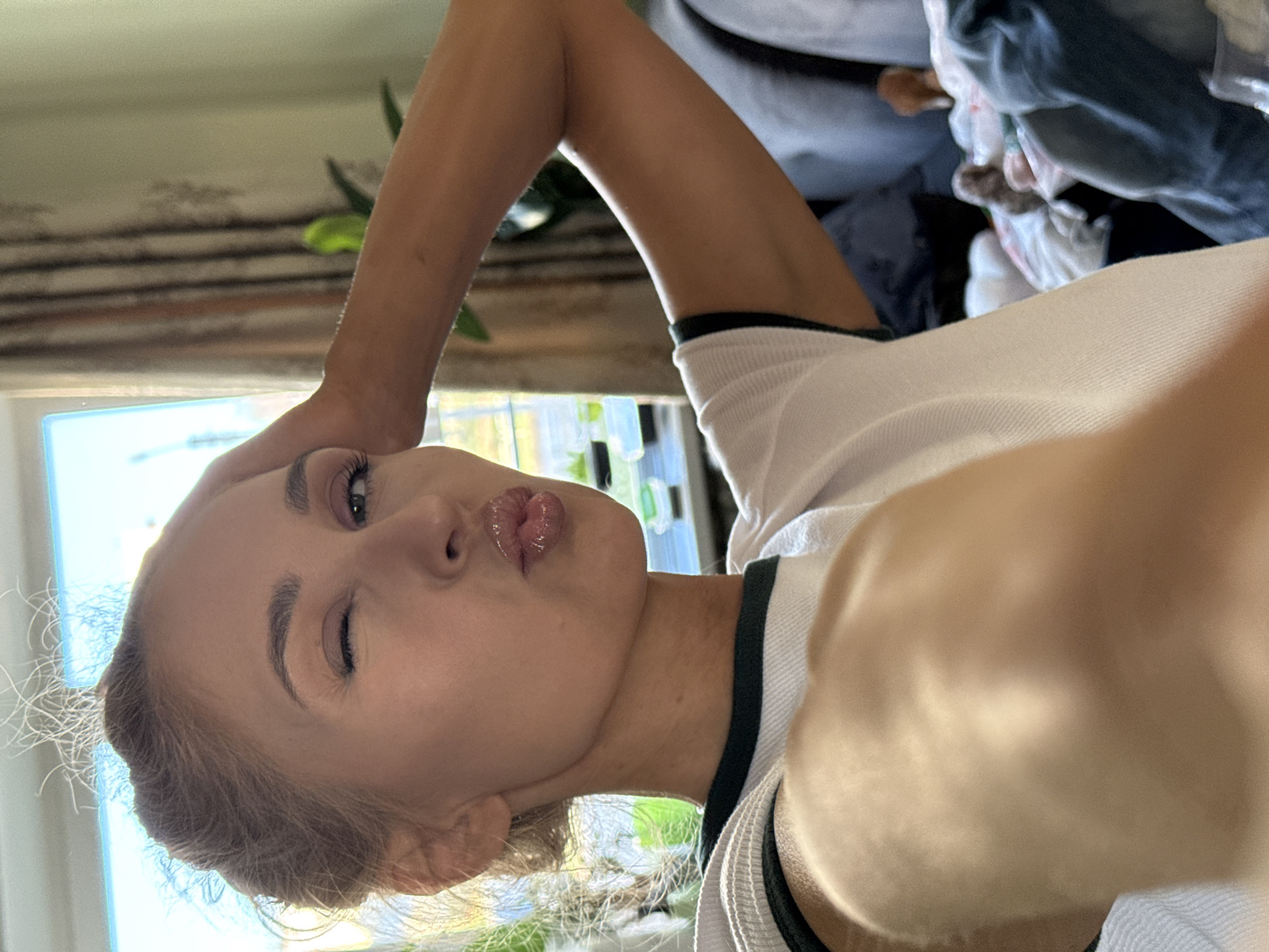 A young woman is the central subject of this close-up, rotated selfie, captured indoors in what appears to be a room in Chaikovsky, Russia. She is shown from the chest up, with her head tilted sideways. The woman has light-colored hair, which is styled away from her face, revealing her features.
She is engaged in the activity of taking a selfie, evident by the angle and her raised arm, likely holding the camera. Her facial expression is playful: she has one eye closed in a distinct wink, and her lips are pursed into a 'kiss' or 'duck face', enhanced by a glossy finish. She appears to be wearing eye makeup, including eyeliner and mascara.
She is dressed in a white top featuring a contrasting dark trim around the neckline, possibly black or dark green. Her right arm is partially visible in the foreground, reaching up to frame the shot.
The background elements are largely out of focus. Bright natural light streams through a window, indicating it is daytime. Beyond the window, some indistinct outdoor greenery or structures can be vaguely seen. Other blurred elements in the room include what appears to be furniture, pillows, or folded clothes in muted colors. There is no visible text in the image.