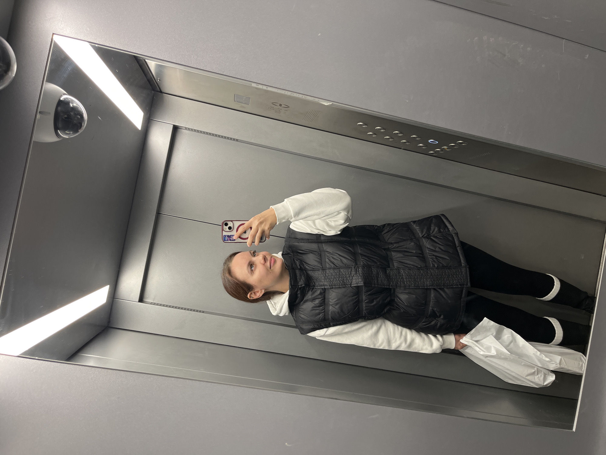 A young woman is taking a selfie in an elevator with a mirrored wall. She is wearing a white hoodie, black puffer vest, and black leggings with white stripes around the ankles. She is holding a white shopping bag. The elevator is gray metal with button controls visible at the top. A security camera is visible in the top corner of the mirror. It is not possible to determine the exact location within Chaikovsky, Russia.