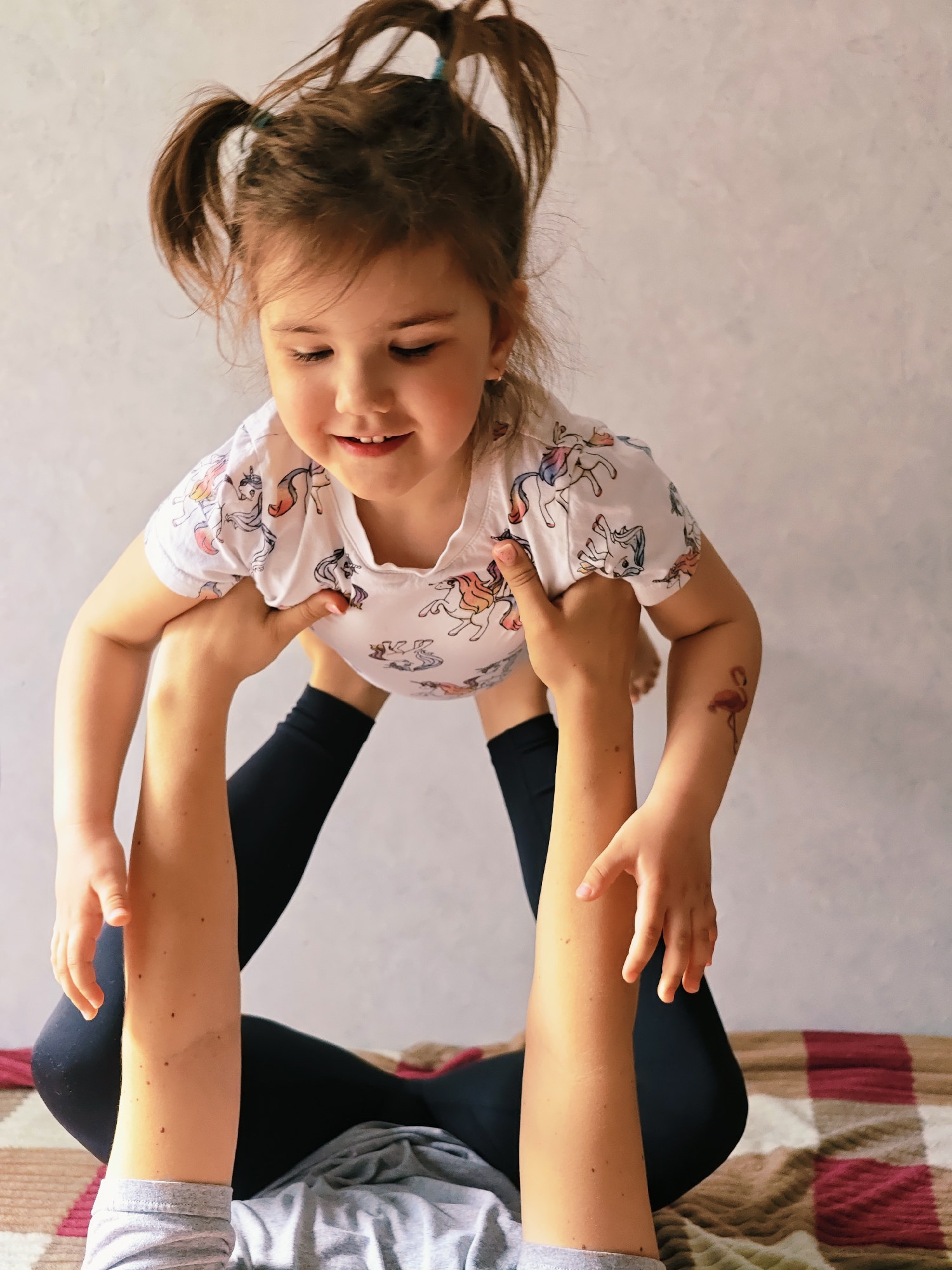 A young girl in a unicorn-printed white t-shirt is being held aloft by an adult. The adult is sitting on a striped blanket wearing black leggings and a gray top. The girl has brown hair in pigtails and a flamingo tattoo on her right arm. The background is a plain light wall. This photo appears to be taken indoors. The city of Chaikovsky, Russia, is not discernible from the visual data.