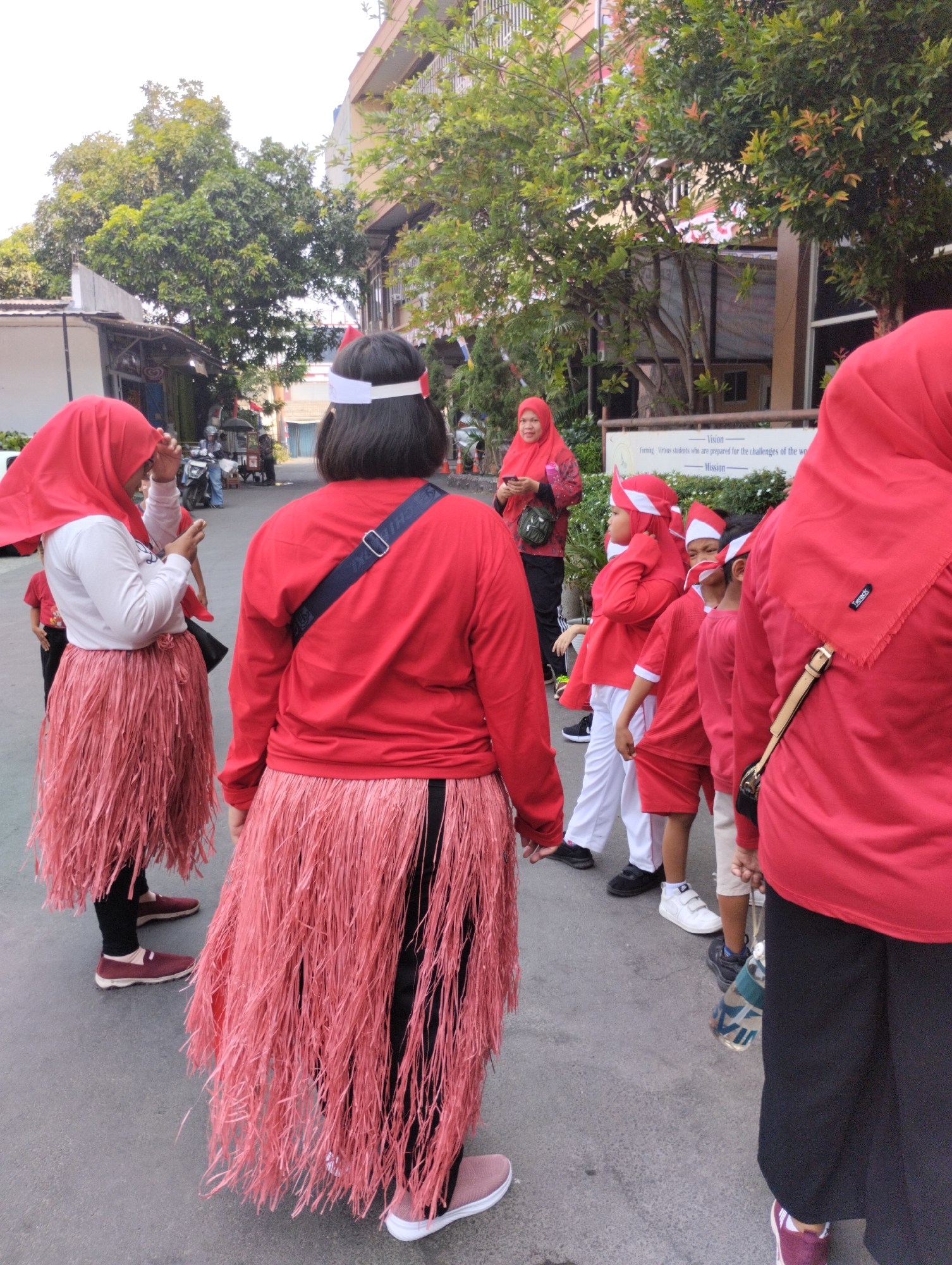 In the city of Bekasi, Indonesia, a group of people, primarily children and a few adults, are seen dressed in matching red attire, participating in an outdoor event or parade. The children are wearing red shirts, red headbands resembling crowns, and some have white headbands. Two individuals, likely adults, are wearing red long-sleeved shirts and pink grass skirts, possibly as part of a costume. One adult in the background is wearing a red hijab and a patterned outfit. They are standing on a paved surface, possibly a street or pathway, with buildings and trees visible in the background. A sign with text is also present on a building. The overall atmosphere suggests a festive or celebratory occasion.