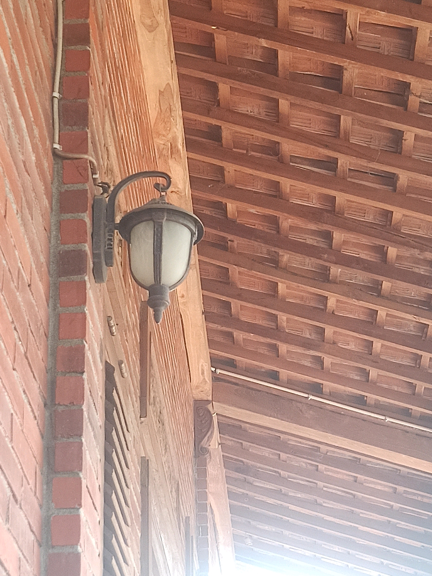 An exterior shot captures a wall mounted light fixture on a brick wall in Bangilan, Indonesia. The light fixture, which appears to be made of dark metal with a frosted glass shade, is attached to the left side of the image. A white conduit runs vertically along the brick wall, to the left of the light fixture. The brick wall itself is made of reddish-brown bricks with gray mortar. To the right of the brick wall, a wooden structure with horizontal beams is visible, suggesting a covered outdoor area or eaves. The lighting conditions suggest it is daytime, with sunlight illuminating the scene.