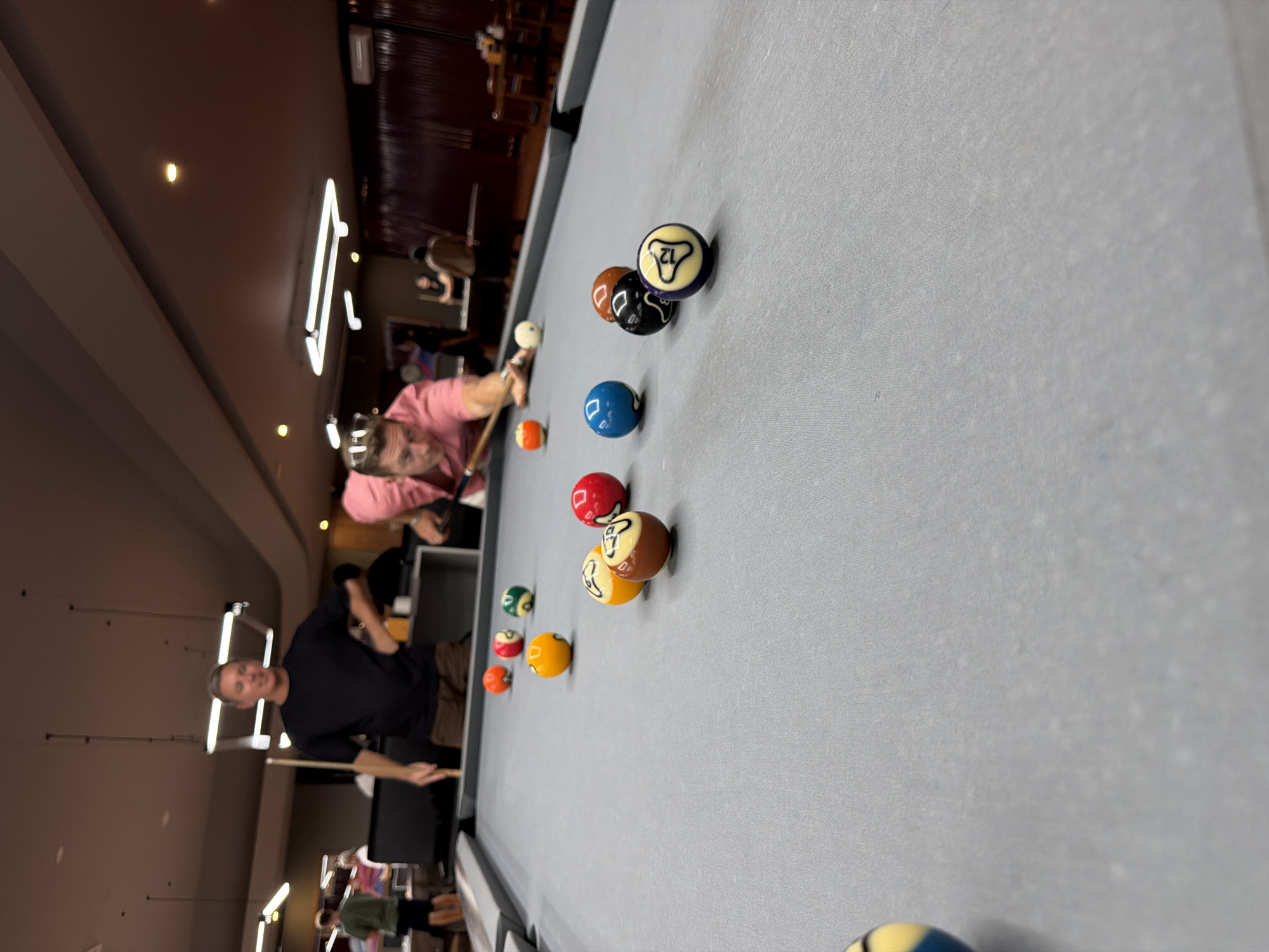 A man in a pink shirt is playing pool in an indoor billiard hall. He is focused on hitting a white cue ball with a cue stick, aiming at a cluster of colored billiard balls on a grey felt table. Several billiard balls are scattered on the table, including solid colors and striped balls. In the background, another man in a black t-shirt watches, holding a cue stick. Other individuals are visible in the background, blurred and out of focus, suggesting a social gathering or a public establishment. The lighting appears to be artificial, with overhead fixtures illuminating the scene. The environment suggests a casual, recreational setting.