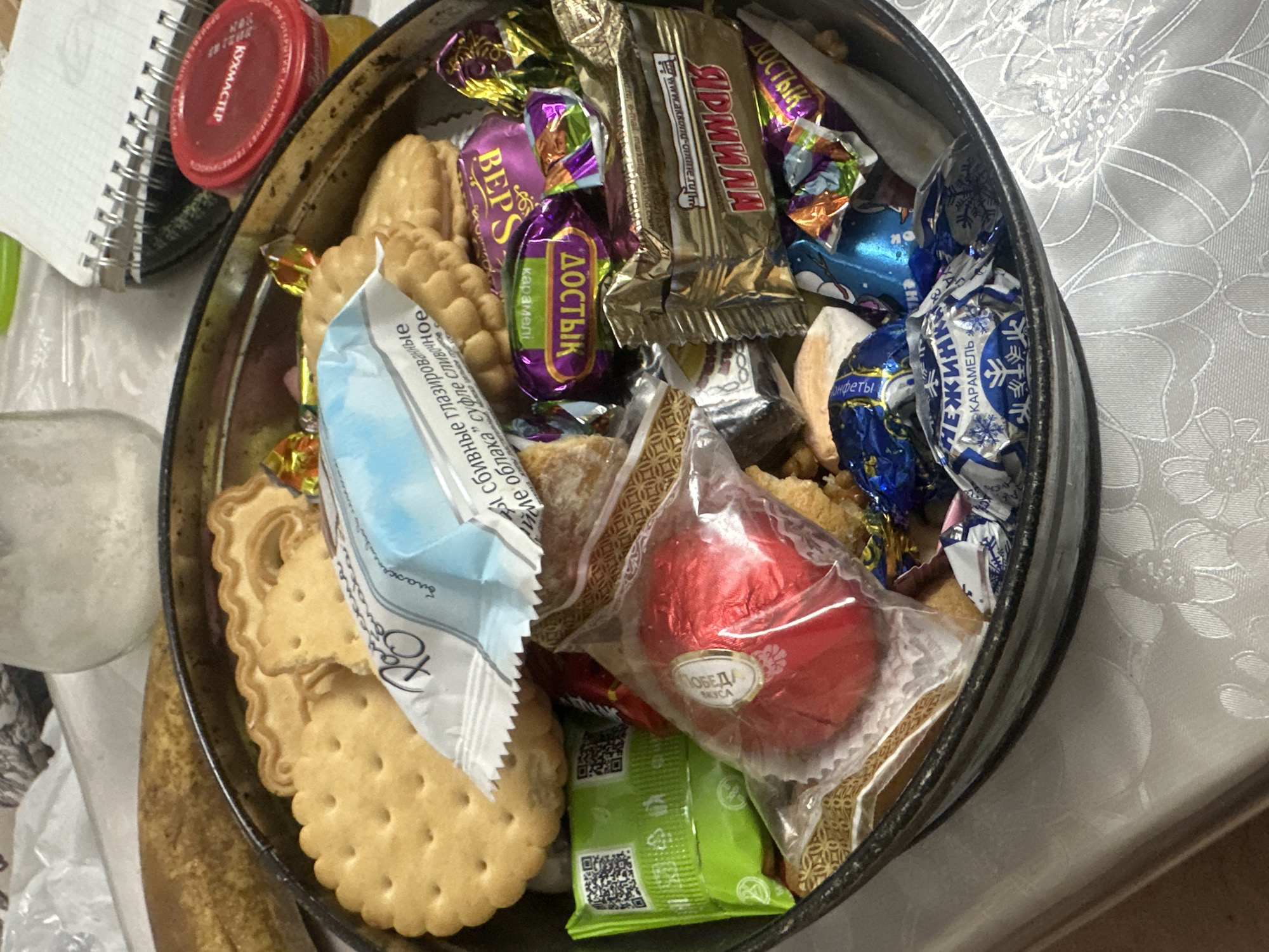 A close-up, overhead shot shows a metal tin filled with various sweets, cookies, and a banana in the upper left corner. The tin, which appears to be old and slightly tarnished, sits on a white tablecloth with a floral pattern. Among the contents are several cream-filled cookies, individually wrapped chocolates with colorful foil, and a shiny red foil-wrapped candy in the center. A torn blue and white wrapper is also visible, displaying Cyrillic text. To the right of the tin, a red-lidded jar and a spiral-bound notebook are partially visible on a table. The lighting is diffused, suggesting an indoor setting. The scene is likely located in a home in Загородный, Russia, based on the presence of Russian text on the wrappers and the typical contents of such a tin.