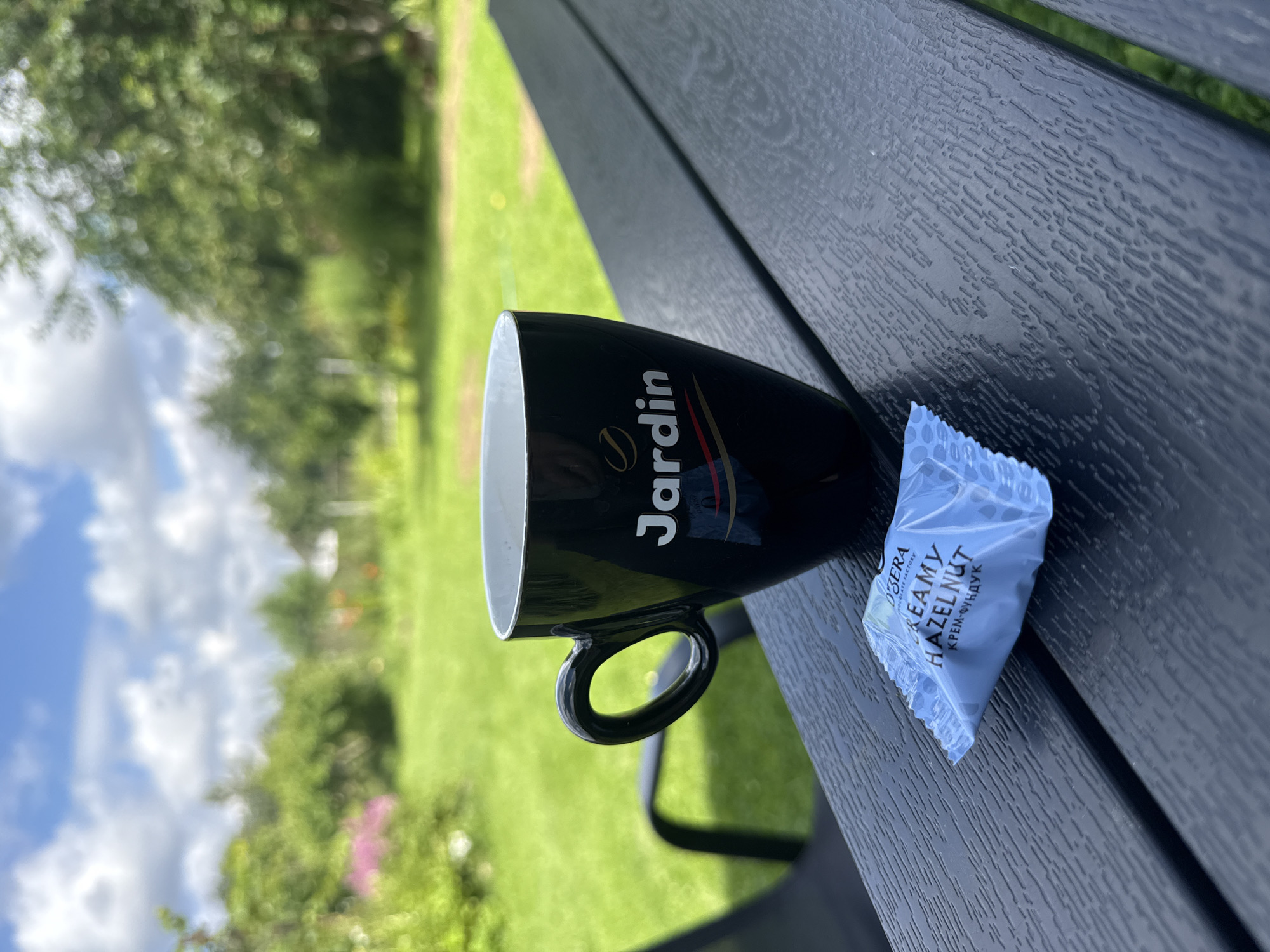 A black mug with the "Jardin" logo and a coffee bean emblem sits on a dark wooden table outdoors. In front of the mug is a small, light blue wrapper with text that reads "OZERO CHOCOLATE FACTORY CREAMY HAZELNUT". The background shows a green lawn, trees, and a cloudy blue sky, suggesting a backyard setting. The image appears to be taken from a slightly elevated angle. No people are visible in the image. The scene does not provide any specific clues to pinpoint its location within Mahilyow, Belarus.