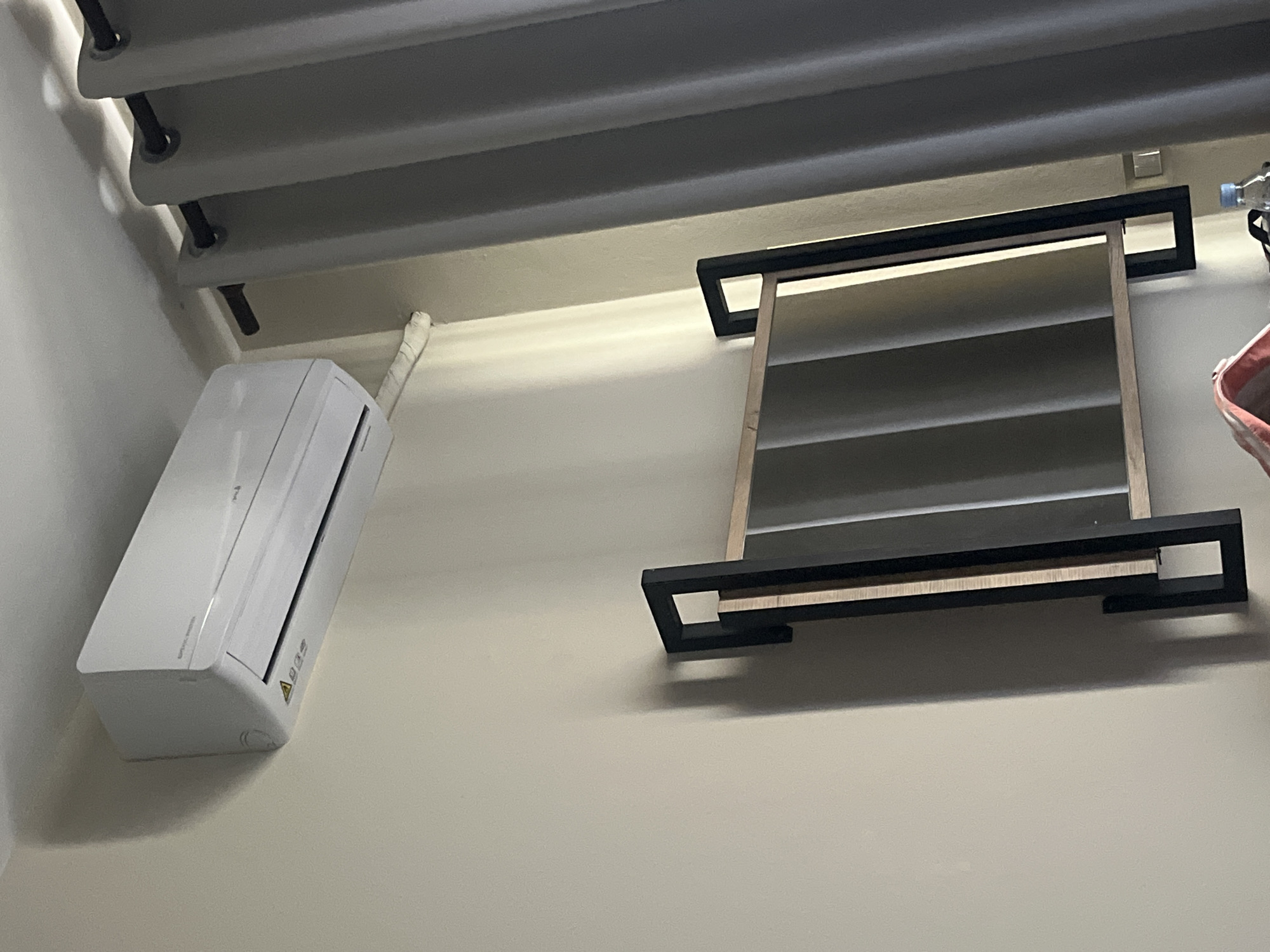 The image depicts an indoor scene, likely a room in a building. The walls are painted in a neutral, off-white color. Visible architectural elements include a layered structure of horizontal, grey-toned bars, possibly part of a ceiling or framework, with black supports. An air conditioning unit, white in color, is mounted on the wall on the left. Next to it there is a duct or tube connecting to the air conditioning unit. A black-framed rectangular structure with horizontal, reflective bars hangs on the wall. The overall environment suggests an interior space with functional appliances and structural elements.