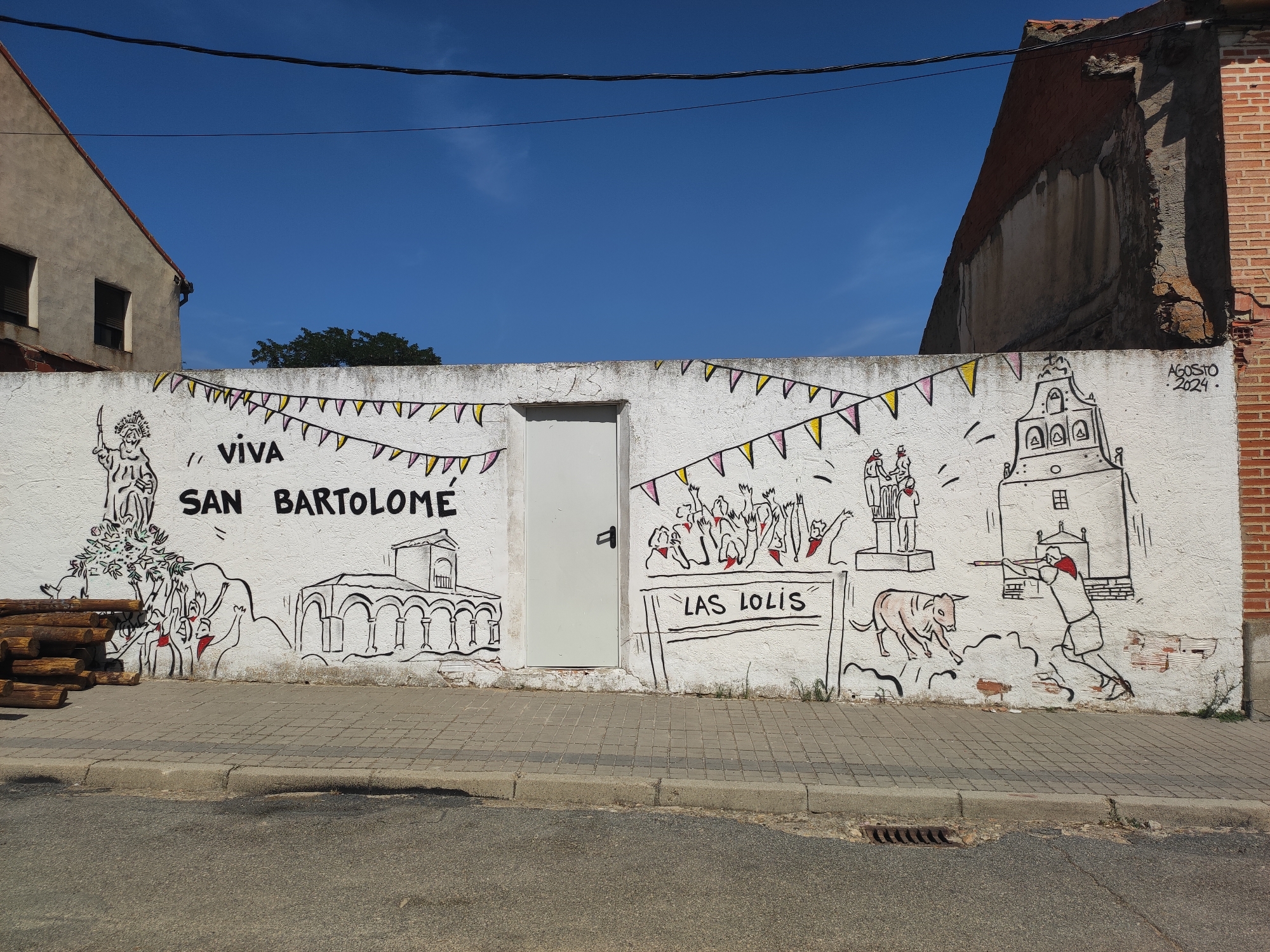 The image shows a mural on a white stucco wall in Sangarcía, Spain. The mural is a cartoon-style depiction of a festive scene, likely related to the San Bartolomé festival. The text "VIVA SAN BARTOLOME" is prominently displayed in black lettering.
On the left side of the mural, a figure resembling a statue or a historical person is depicted standing next to a tree with red accents. Below this, a stack of logs is piled against the wall. To the right of the text, a series of cartoonish figures are shown with raised arms, suggesting celebration. In the center, there is a white door integrated into the wall.
Further to the right, a sign reads "LAS LOLIS." Below this, a pink cartoon bull is shown in motion, with a stick-figure person holding a long pole facing it, seemingly engaged in a bull-related activity. To the far right, a stylized depiction of a church or bell tower is present. Above the mural, festive triangular flags in yellow and pink are strung across the wall.
The date "AGOSTO 2024" is inscribed in the upper right corner of the mural. The scene is set against a clear blue sky. The wall itself shows some signs of wear and tear, with patches of exposed brick visible in places. The mural is situated on a sidewalk, with a street in the foreground. Electrical wires are visible overhead. The surrounding environment includes parts of buildings, one with a red brick facade and another with a textured beige finish.