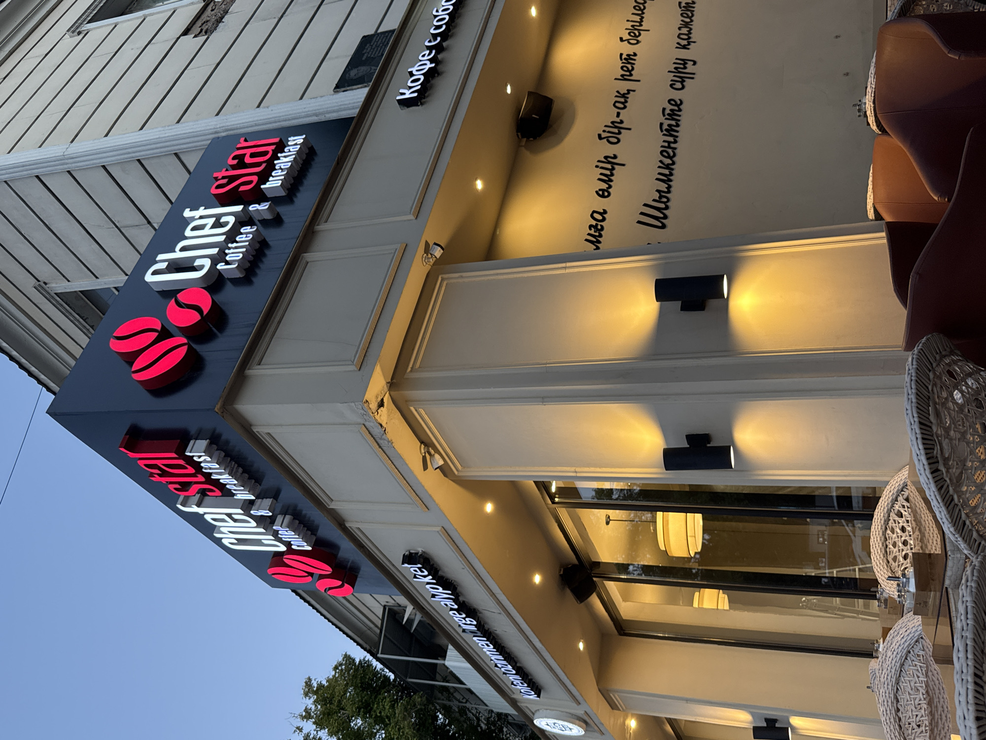The image shows the exterior of a cafe named "Chef Star" with the slogan "Coffee & Breakfast" visible. The cafe's signage features the name "Chef Star" in prominent white and red lettering, accompanied by graphics of red coffee beans. Below this main sign, there is another sign with text in what appears to be Kazakh or a similar language, reading "Kafeni özíńmen urge alyp kel" which translates to "Take your coffee with you". Further along the facade, there is a sign that says "Кофе с собой", which means "Coffee to go" in Russian. The cafe is illuminated by warm, yellow outdoor lighting. There are tables and chairs visible outside the cafe, with some chairs made of wicker and others appearing to be plush leather armchairs. The building itself has a textured facade with decorative elements. In the background, a tree is visible. The sky is a clear blue, suggesting it is daytime. The overall scene depicts a street-level view of a commercial establishment in a city setting.