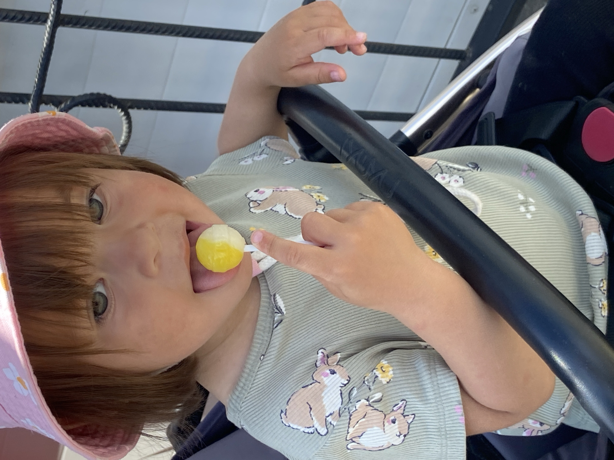 A young child, possibly a toddler, is sitting in a stroller outdoors. The child is wearing a light-colored shirt with a rabbit pattern and a pink sun hat with white flowers. The child has a yellow lollipop in their mouth and is holding the stick with their hand. The stroller has a black handle and a dark-colored seat with a red button visible. The background shows a metal fence or railing and a light-colored surface, possibly a wall. The photo was taken in Tyumen, Russia.