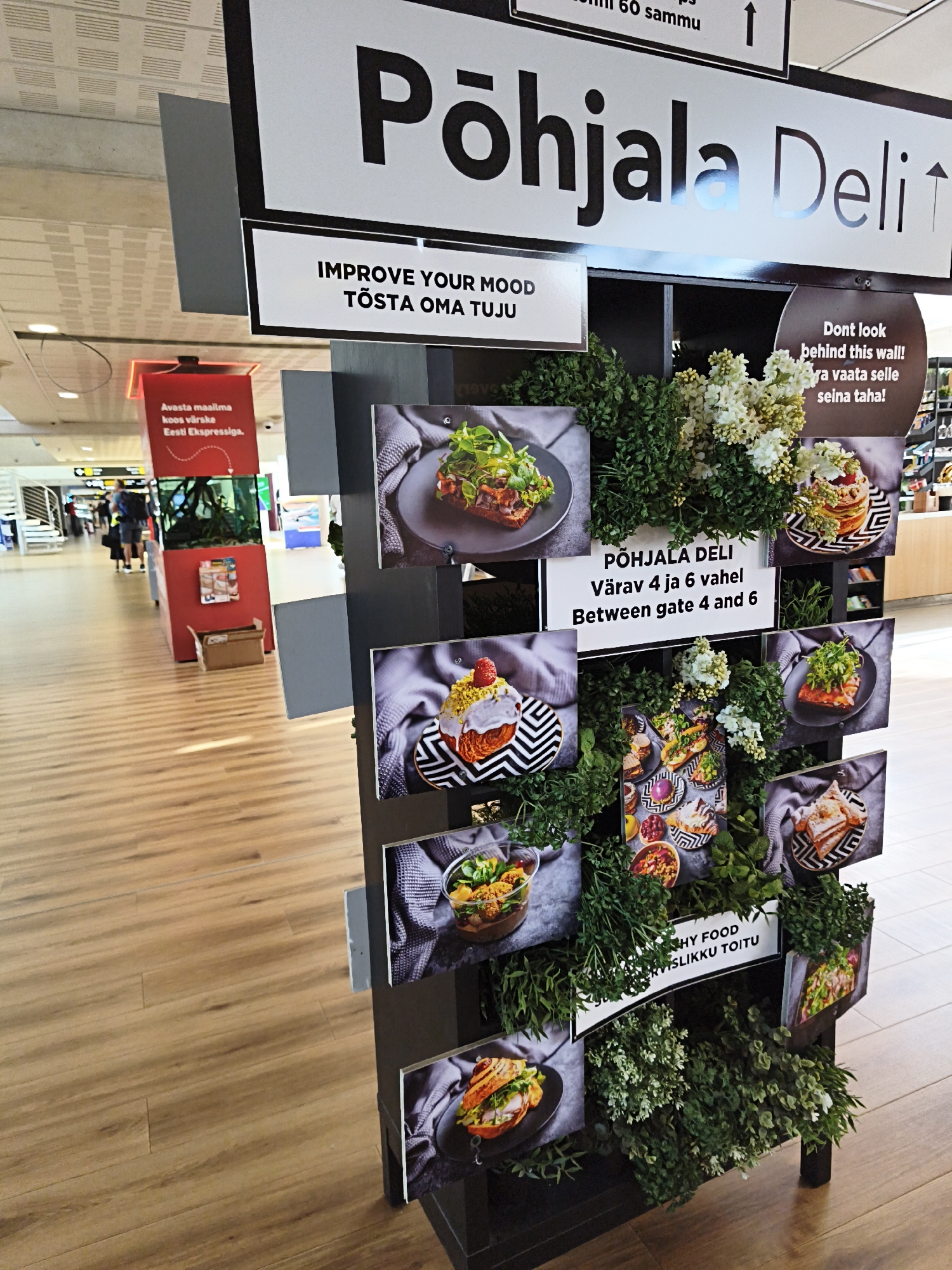 The image shows a display for "Põhjala Deli" at Tallinn Airport, Estonia. The display features images of food and greenery, with text in English and Estonian. The text "IMPROVE YOUR MOOD TÕSTA OMA TUJU" is prominently displayed. Another sign reads "PÕHJALA DELI VÄRAV 4 JA 6 VAHEL BETWEEN GATE 4 AND 6". A third sign states "DONT LOOK BEHIND THIS WALL! ÄRA VAATA SELLE SEINA TAHA!". In the background, a red pillar with a sign about "Eesti Ekspressiga" and an aquarium is visible, along with people walking in the airport concourse. The overall environment is a modern airport interior with wooden flooring.