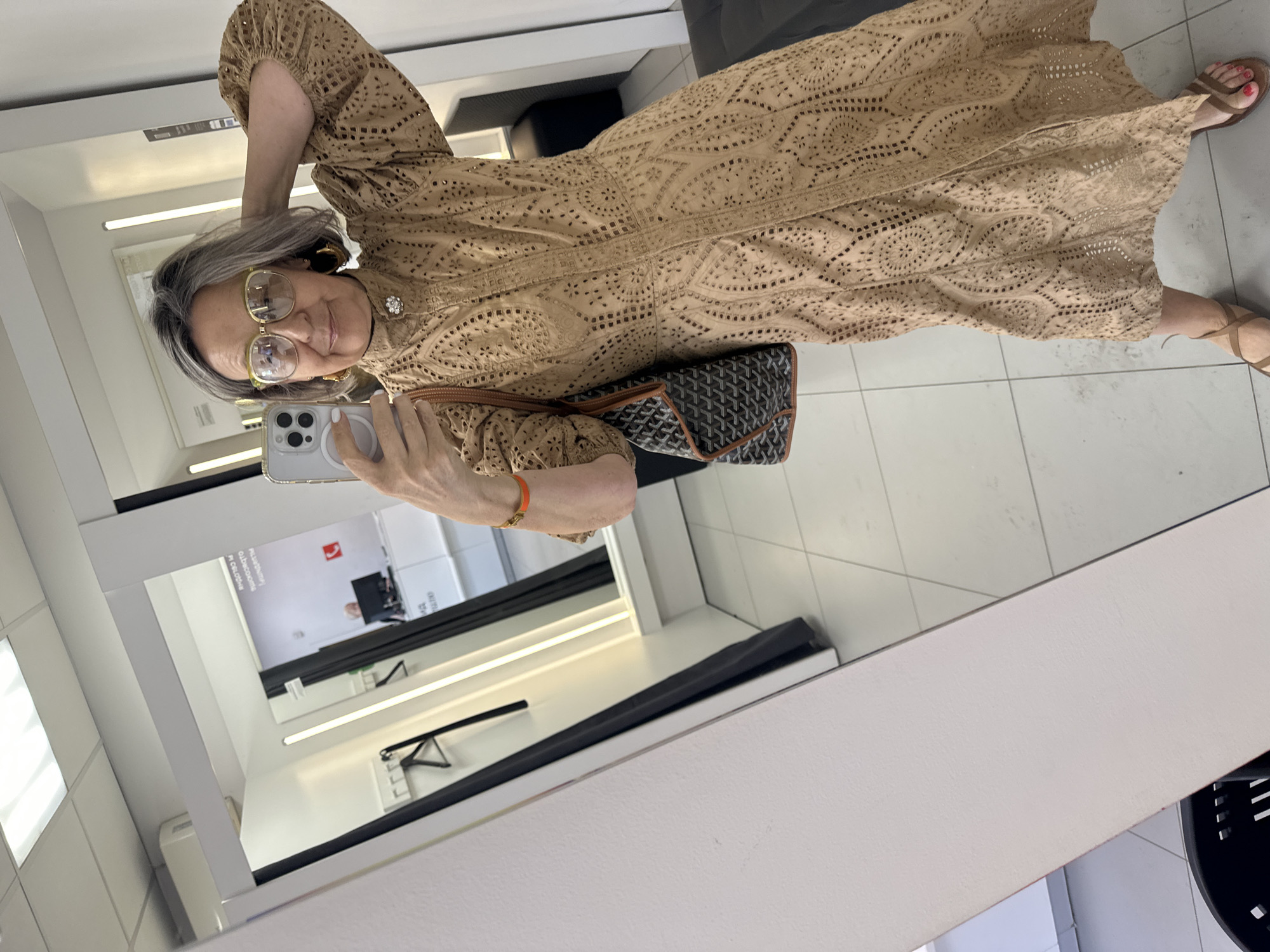 A woman with gray hair and glasses is taking a selfie in a mirror in what appears to be a fitting room or store. She is wearing a tan, eyelet dress and carrying a Goyard tote bag. She has on hoop earrings and an orange bracelet. The fitting room has bright lighting and a tiled floor. There are clothes racks and a black bench visible in the reflection.