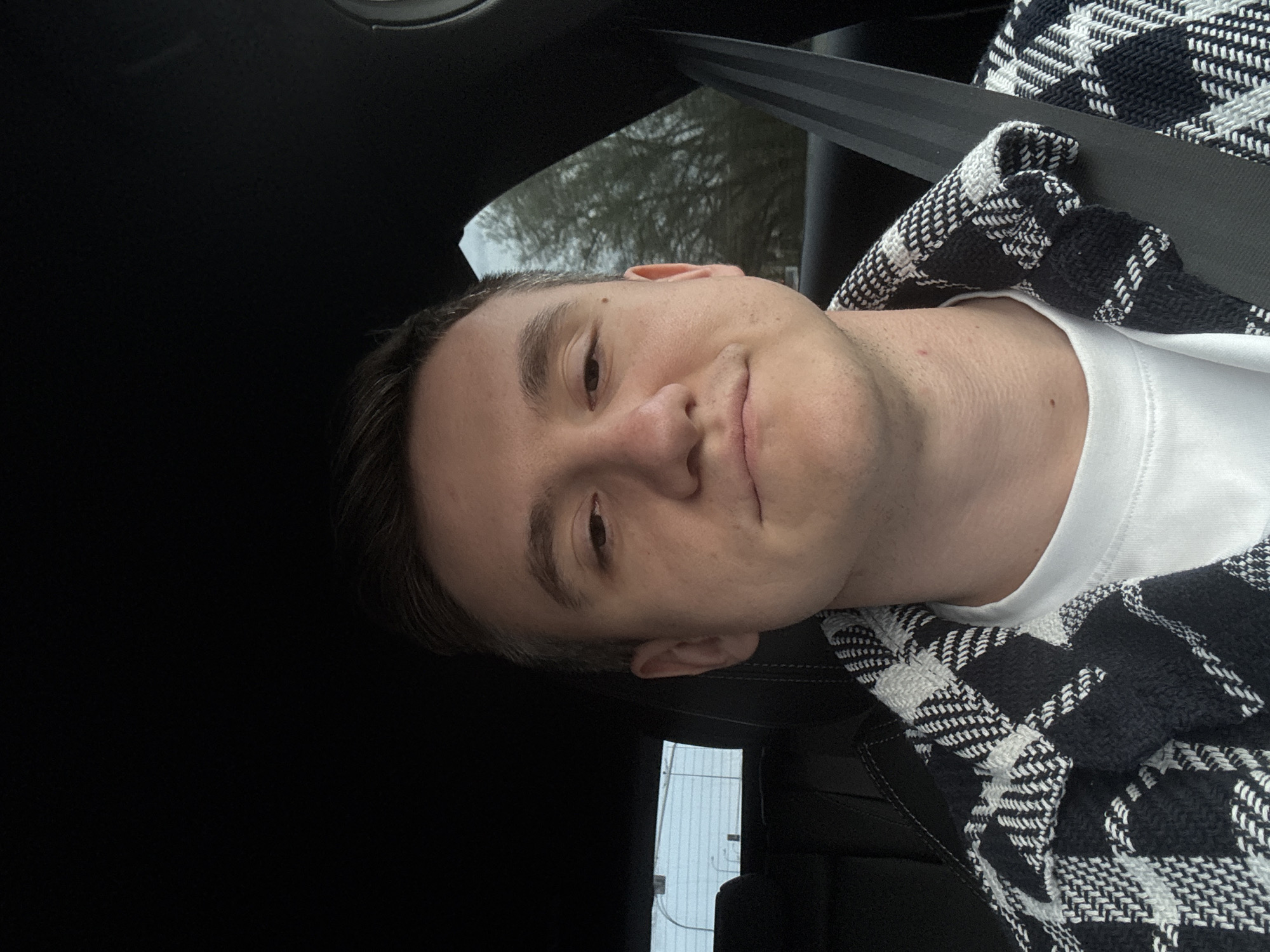 A selfie-style portrait of a Caucasian male in a car. He is wearing a white t-shirt and a black and white patterned jacket. A seatbelt crosses his chest. Through the car windows, trees are visible, and a building with white siding and vertical lines is partially seen. The interior of the car is mostly black. The location cannot be determined to be Warsaw, Poland from the visual information in the image.