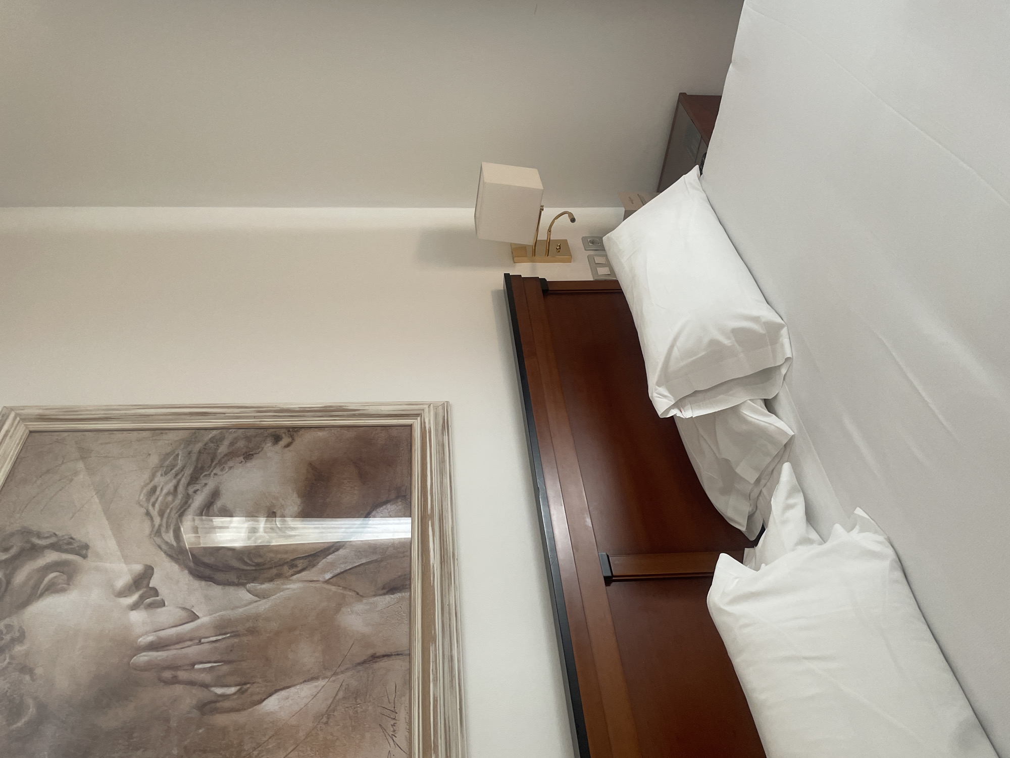 The image shows a close-up view of a bed with white linens and pillows. A wooden headboard is visible behind the pillows. Above the headboard, a framed artwork depicting two figures, rendered in a muted color palette, hangs on a white wall. To the right of the bed, a bedside table with a lamp is partially visible. The scene appears to be a hotel room in Sevilla, Spain, judging by the style and the presence of what might be a hotel information card on the bedside table.