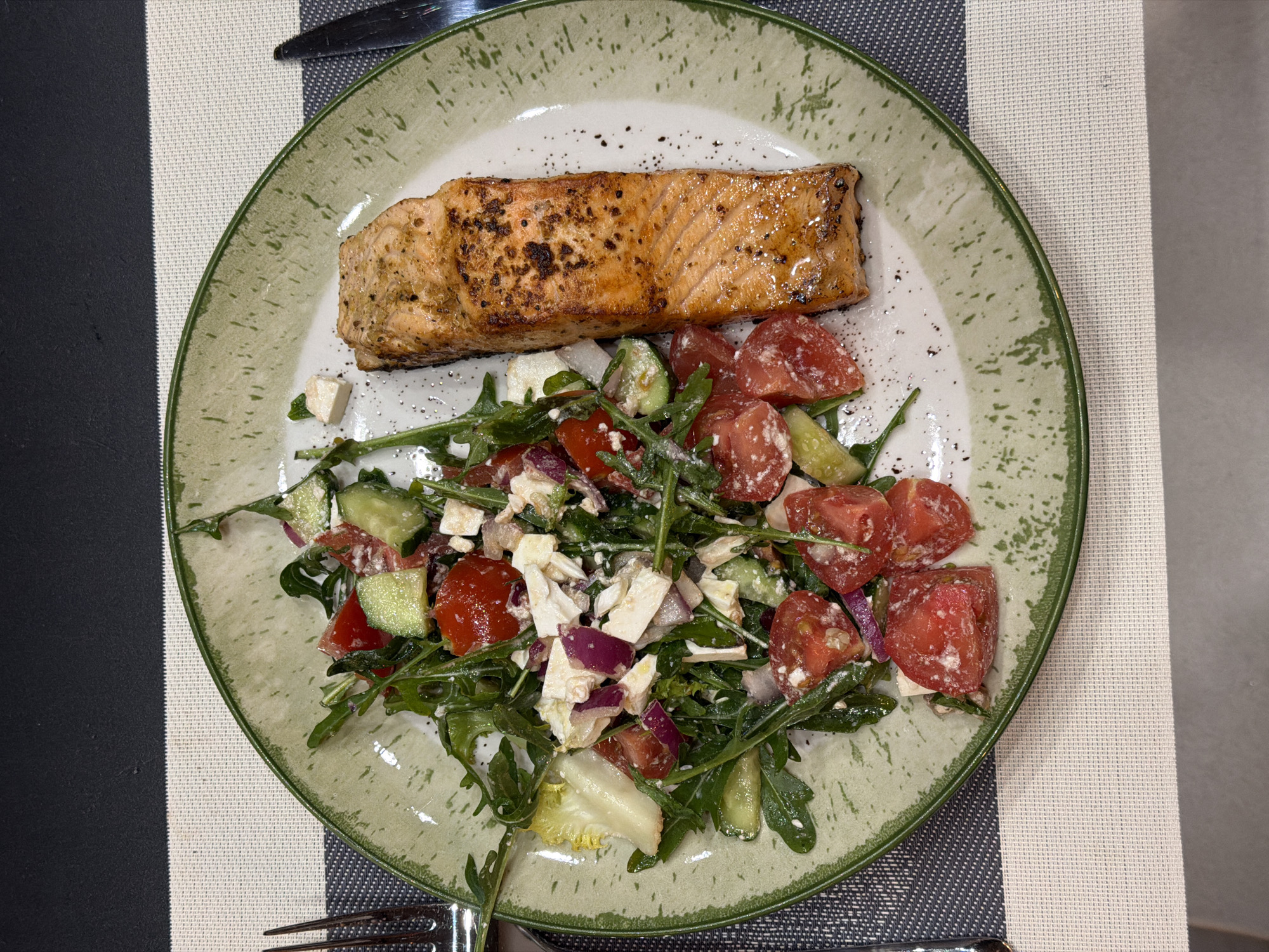 A plate of food is centered in the frame. The plate has a pale green mottled surface with a darker green rim. On the plate is a piece of cooked salmon and a salad made of chopped tomatoes, cucumbers, red onion, feta cheese, and arugula. Black pepper is sprinkled over the salmon and salad. The plate sits on a placemat with alternating gray and white stripes. A fork and knife flank the plate. The location is most likely a restaurant or a private residence, but it is not possible to determine it to be Ryazan, Russia, from the image.