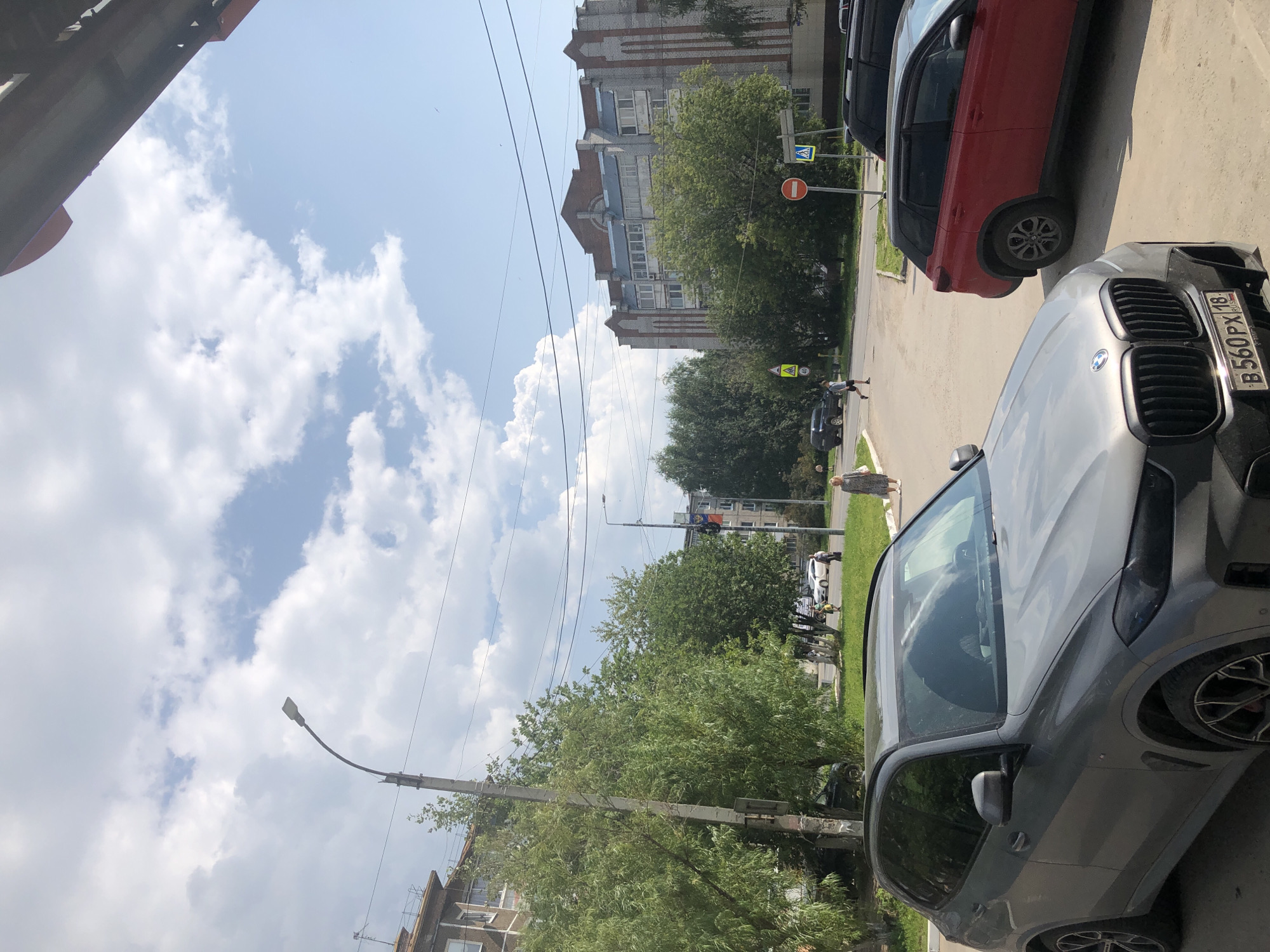 The image depicts a street scene in Solikamsk, Russia. In the foreground, a grey BMW SUV is parked, partially obscuring the view of a red SUV behind it. The street is paved, and a sidewalk is visible. Several trees line the street, with lush green foliage. In the background, a multi-story apartment building with light-colored brickwork and red decorative elements stands prominently. Another building, also appearing to be an apartment complex, is visible further down the street. Several road signs are present, including a yield sign and a speed limit sign. A few pedestrians can be seen walking on the sidewalk and crossing the street. The sky is partly cloudy with white cumulus clouds against a blue background. A street lamp is visible on the left side of the image.