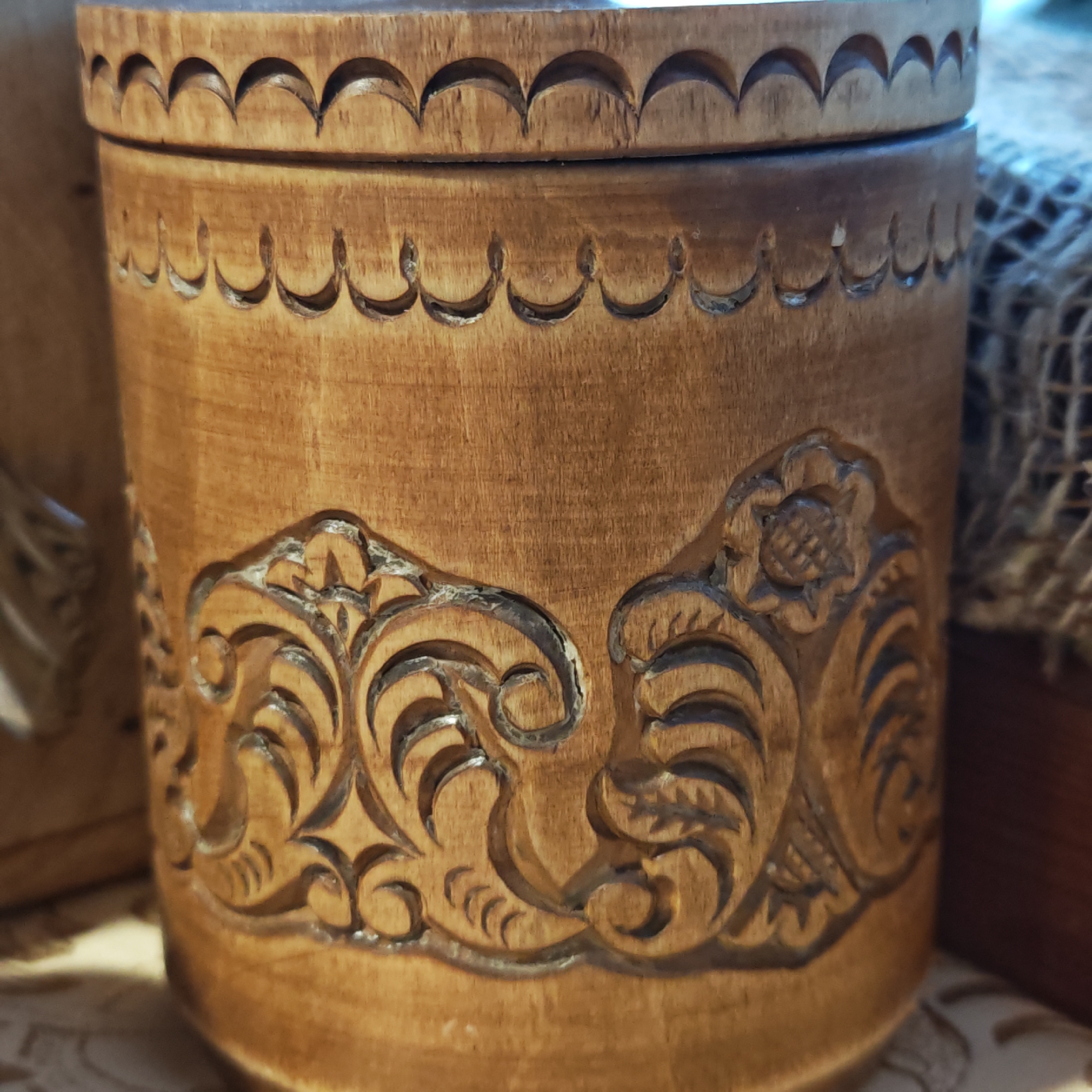 The image depicts a cylindrical wooden container with a lid. The container is light brown in color and features carved decorative patterns. The patterns include a floral motif near the top, a row of semicircular carvings around the lid's edge, and a complex design with leaves and curls covering the lower portion of the container. The image was taken in Rostov, Russia.