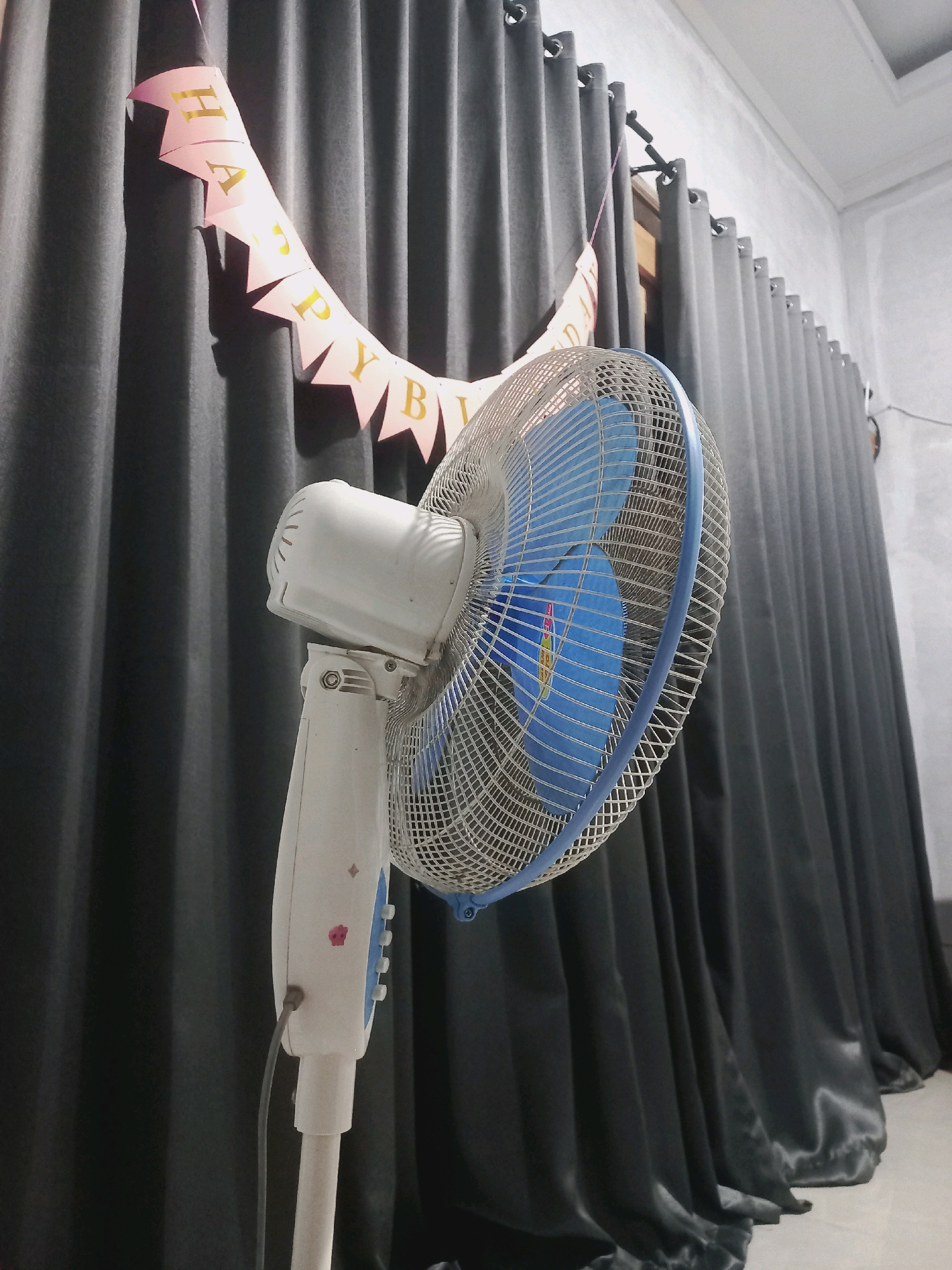A white pedestal fan with a blue fan blade sits in front of a dark grey curtain. A pink and gold "Happy Birthday" banner is draped across the curtain behind the fan. The scene appears to be indoors, likely a room in a house in Gambiranom, Indonesia, possibly decorated for a celebration.