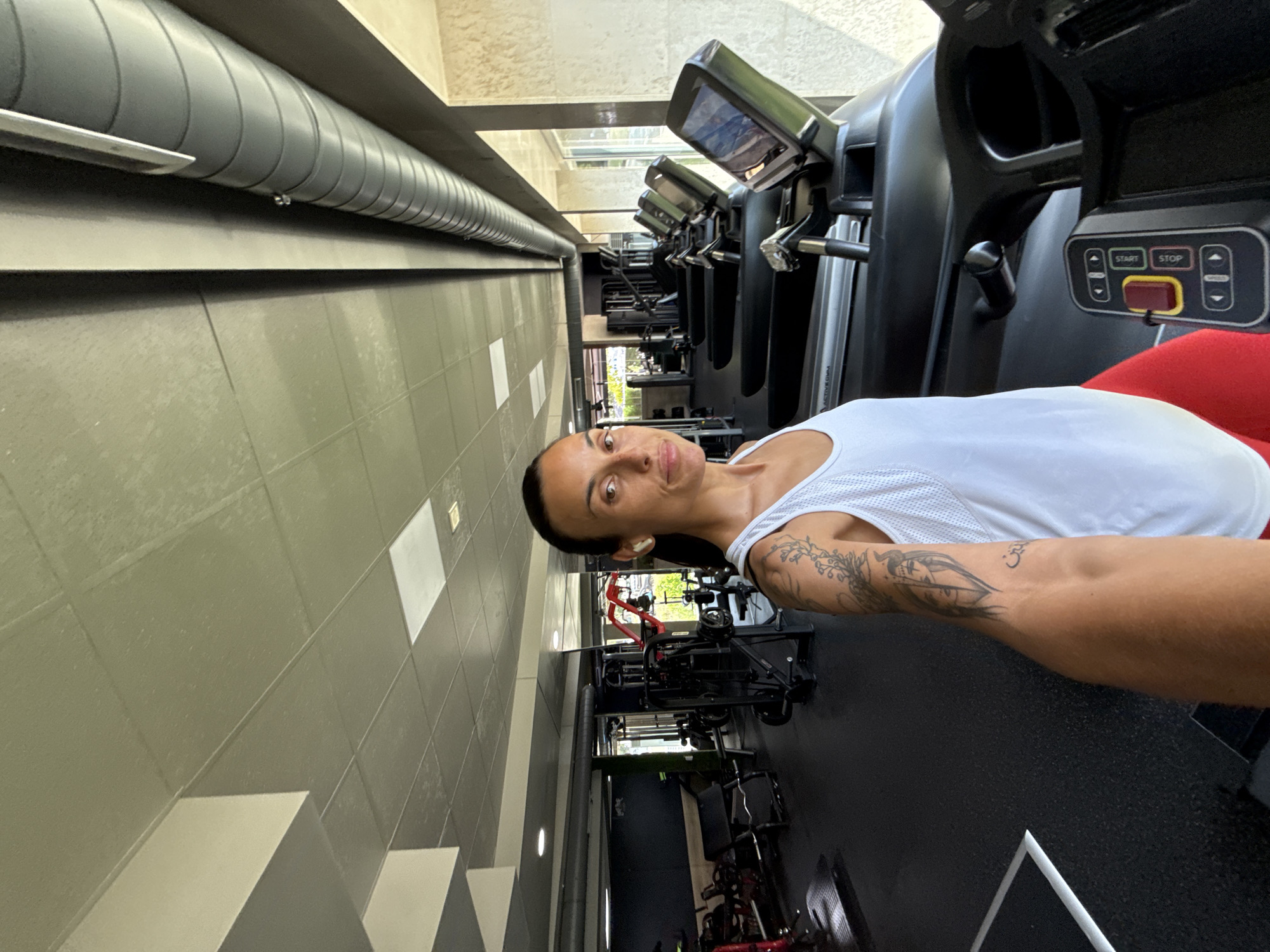 A woman with dark hair pulled back and visible tattoos on her arm is taking a selfie in a gym. She is wearing a white tank top and red leggings. She is standing in front of a row of treadmills, with the machines and other gym equipment visible in the background. The gym has a modern industrial design with a grey tiled ceiling and exposed ductwork. Large windows on the right side of the frame allow natural light to enter the space. The image does not provide any specific information about the location in the city of Pleven, Bulgaria.