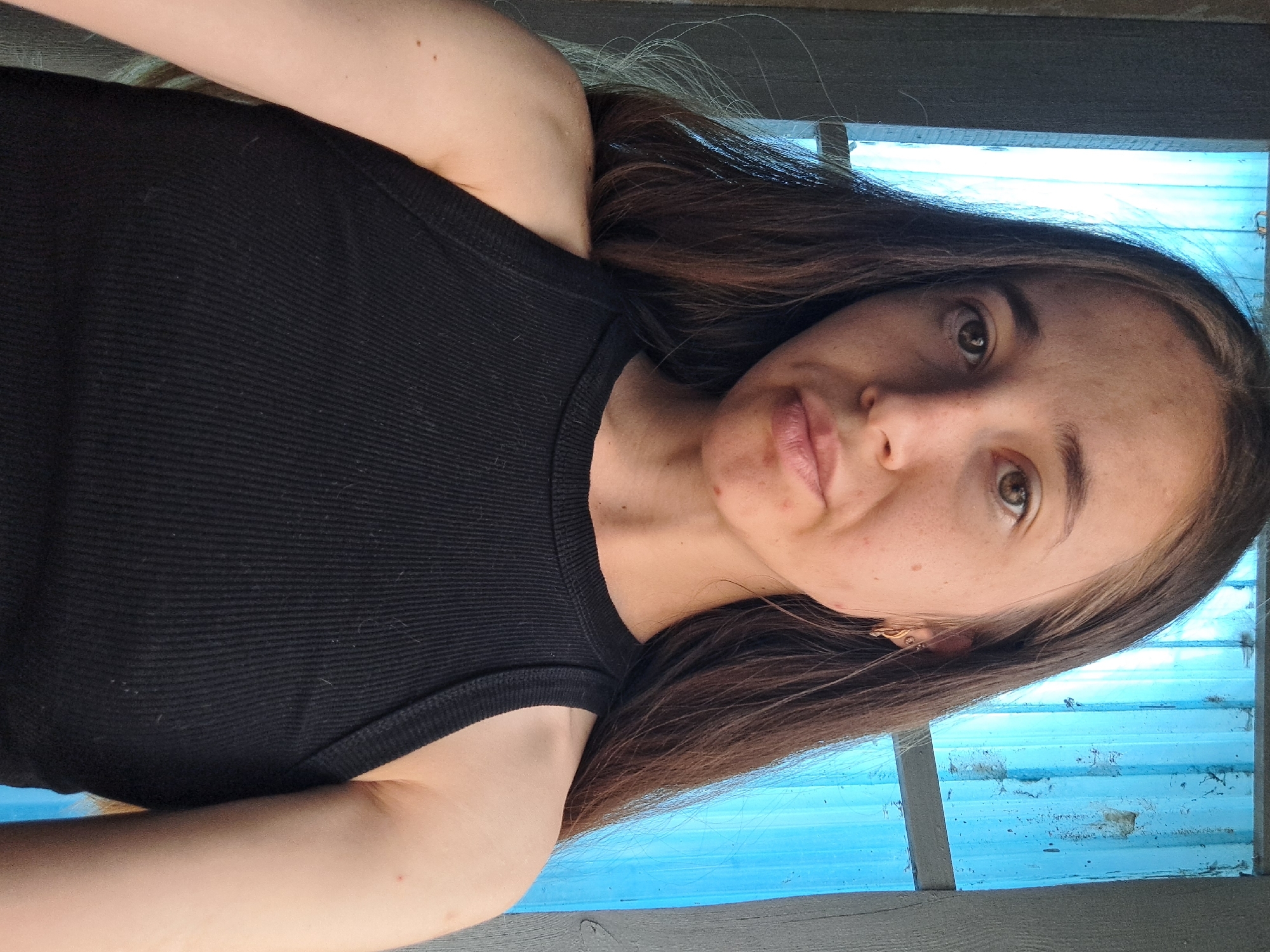 A young woman with long brown hair is posing for a selfie. She is wearing a black ribbed tank top. Her head is tilted slightly to her left. The background consists of a window with blue vertical panes, possibly corrugated plastic, and a wooden frame. There is also some evidence of wear and tear on the blue panes. The lighting suggests it is daytime, possibly outdoors or in a well-lit interior. The image appears to have been taken in the village of Vypasne, Ukraine.