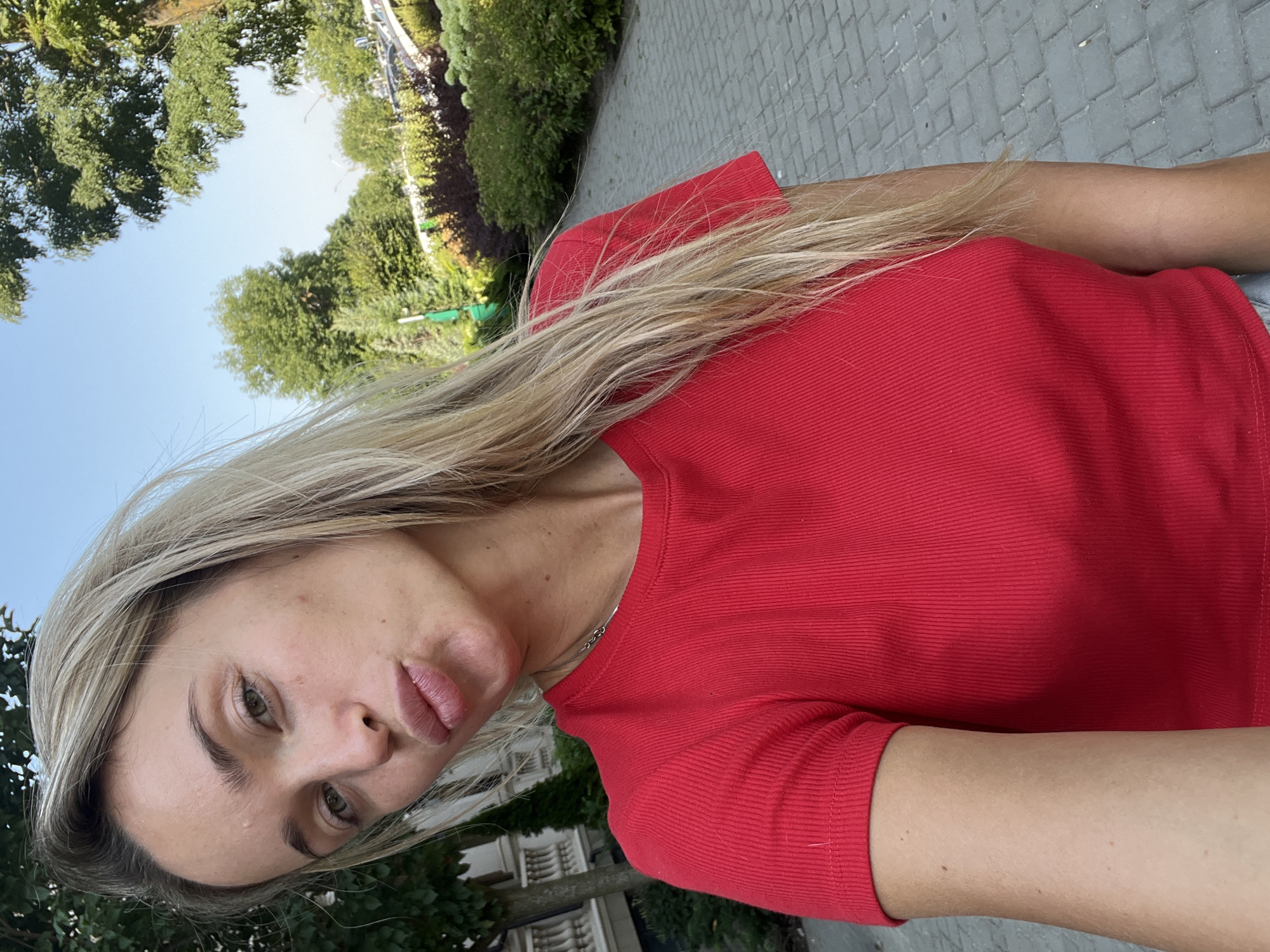 In a selfie taken outdoors, a young blonde woman wearing a red ribbed t-shirt puckers her lips towards the camera. She is standing on a paved walkway, with a manicured garden to her right and a building with decorative balustrades to her left. Lush green trees and a clear blue sky form the background. The overall impression is that of a casual outdoor moment in a well-maintained urban or suburban setting.