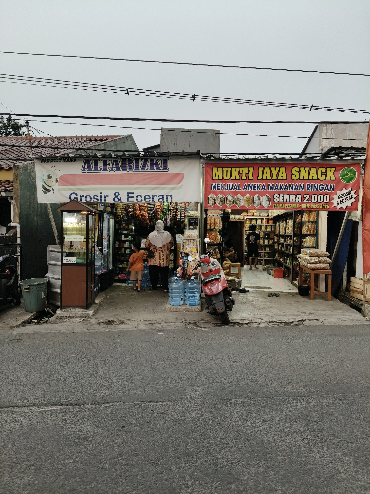 The image captures a storefront in South Tangerang, Indonesia, featuring two adjacent small businesses. The left side is marked with a banner that reads "ALFARIZKI Grosir & Eceran," suggesting it's a wholesale and retail store. A cartoon bee illustration is present on this banner. In front of this store, a display case with bottled drinks and a price tag of "12.000" is visible. A child in an orange t-shirt and shorts stands near the entrance, while an adult wearing a hijab and a patterned shirt stands behind them, facing into the store. Several large water cooler bottles are stacked outside.
To the right, another store displays a red banner with "MUKTI JAYA SNACK" in yellow lettering. Below this, it states "MENJUAL ANEKA MAKANAN RINGAN" (selling various snacks) and "SERBA 2.000" (all for 2,000). A phone number is also listed. This store appears to specialize in snacks, with shelves packed with packaged goods. Inside, two individuals are visible; one is seated, and another is standing in the background. A red scooter is parked in front of this snack store. The environment appears to be a street-level commercial area with overhead power lines and residential-style roofs visible in the background. The overall scene depicts a typical local market or neighborhood shop setting.