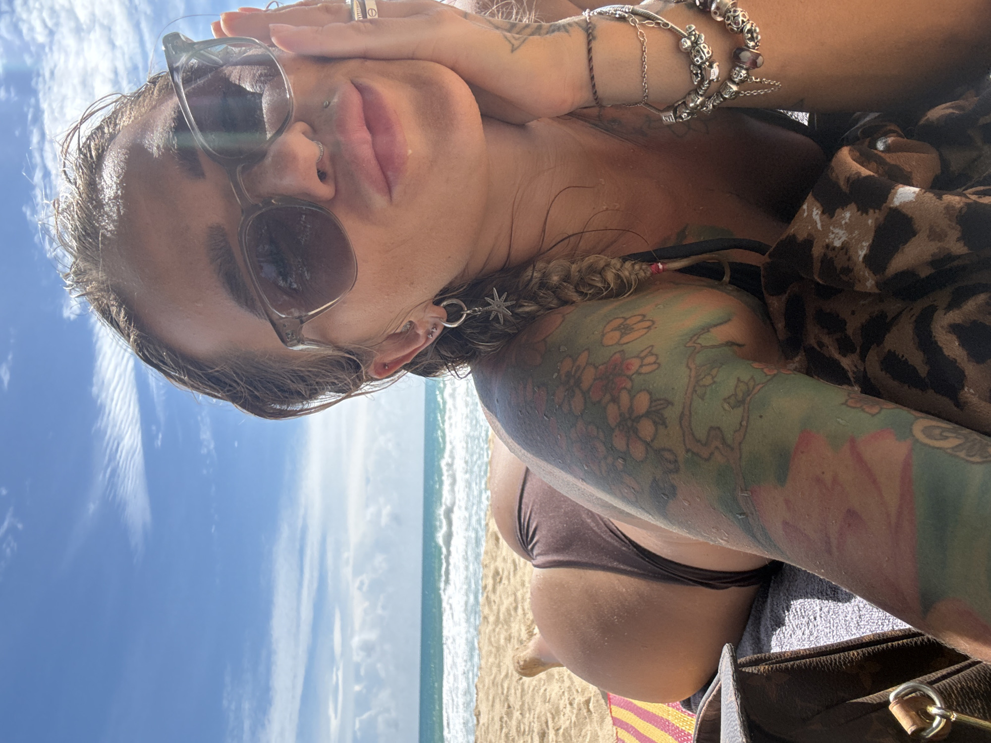 A woman is lying on a beach in Choeng Thale, Thailand, with the ocean and sky in the background. She is wearing sunglasses and a brown bikini. She has tattoos on her arm and is wearing jewelry. The sand is light brown, and there are some colorful stripes on a towel or mat beneath her. The water is blue and there are waves. The sky is blue with some white clouds.