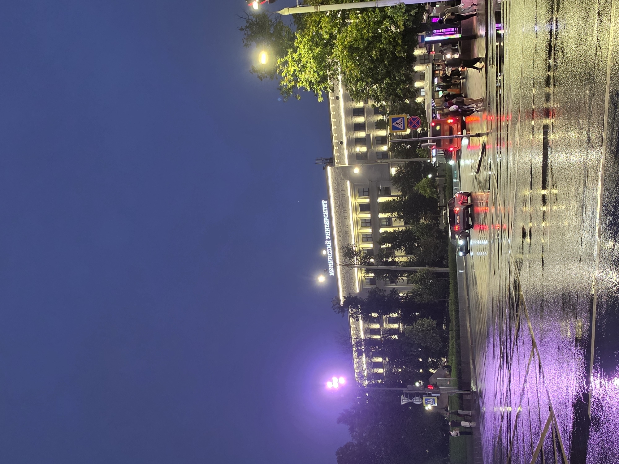 The image depicts a nighttime street scene in Nizhny Novgorod, Russia, featuring a prominent building with illuminated signage that reads "МИНСКИЙ УНИВЕРСИТЕТ" (Minsk University). The architecture of the building is neoclassical, with numerous windows and decorative lighting. The street in the foreground is wet and reflective, indicating recent rain, with purple and yellow light reflections from nearby sources. Several cars, including a red sedan and a dark SUV, are visible on the wet road. A bus is also present, with its taillights illuminated. Pedestrians are seen on the sidewalk and crossing the street. Traffic lights and street signs are also present. Trees line the street and surround the building, their foliage illuminated by streetlights. The sky is dark and cloudy, typical of a night scene.