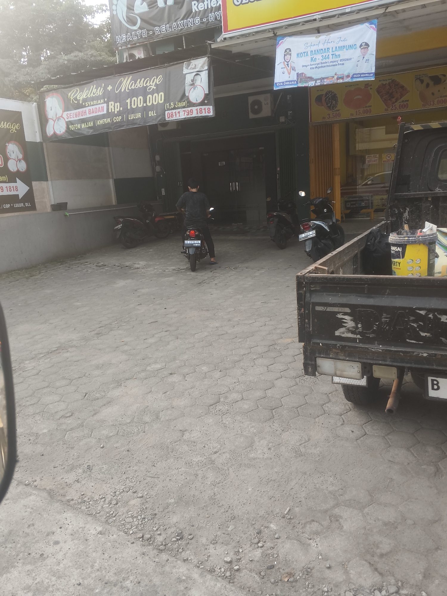 The image shows an exterior view of a building in Bandar Lampung, Indonesia, likely a commercial establishment. In the foreground, the rear of a black pickup truck is visible, with a bucket of yellow paint and some other items in the truck bed. The truck's license plate starts with "B". The ground is paved with hexagonal concrete blocks, with some scattered gravel.
Behind the truck, a person is sitting on a dark-colored motorcycle, facing away from the camera and entering a dark entryway of the building. Several other motorcycles are parked outside the building.
Above the entryway, a banner advertises "Refleksi + Massage SYARIAH" with pricing and a phone number. To the right of this banner, a larger banner displays "SELAMAT HARI JADI KOTA BANDAR LAMPUNG KE - 344 THN". This banner features images of two individuals, possibly local officials, and text related to the city's anniversary. Further to the right, a sign with food items and prices is visible, suggesting the establishment might also offer food or snacks.
The overall environment appears to be a street scene in a commercial area, with overcast skies contributing to diffused lighting.