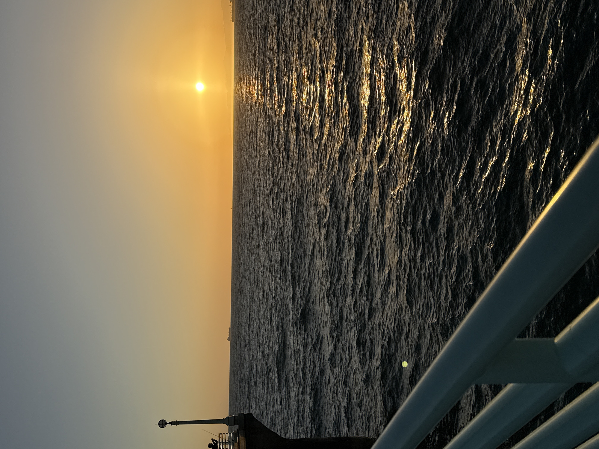 The image depicts a sunset over the Pacific Ocean, viewed from the Manhattan Beach Pier in Manhattan Beach, California. The sun, a bright orb, is positioned in the center-right of the frame, casting a golden-orange glow across the sky and shimmering path on the dark, rippling water. On the left side of the image, a silhouetted figure stands on the pier, holding a fishing rod. A lamppost with a circular light fixture is also visible on the pier. In the distance, on the horizon, several cargo ships can be seen. The foreground features the white railing of the pier.