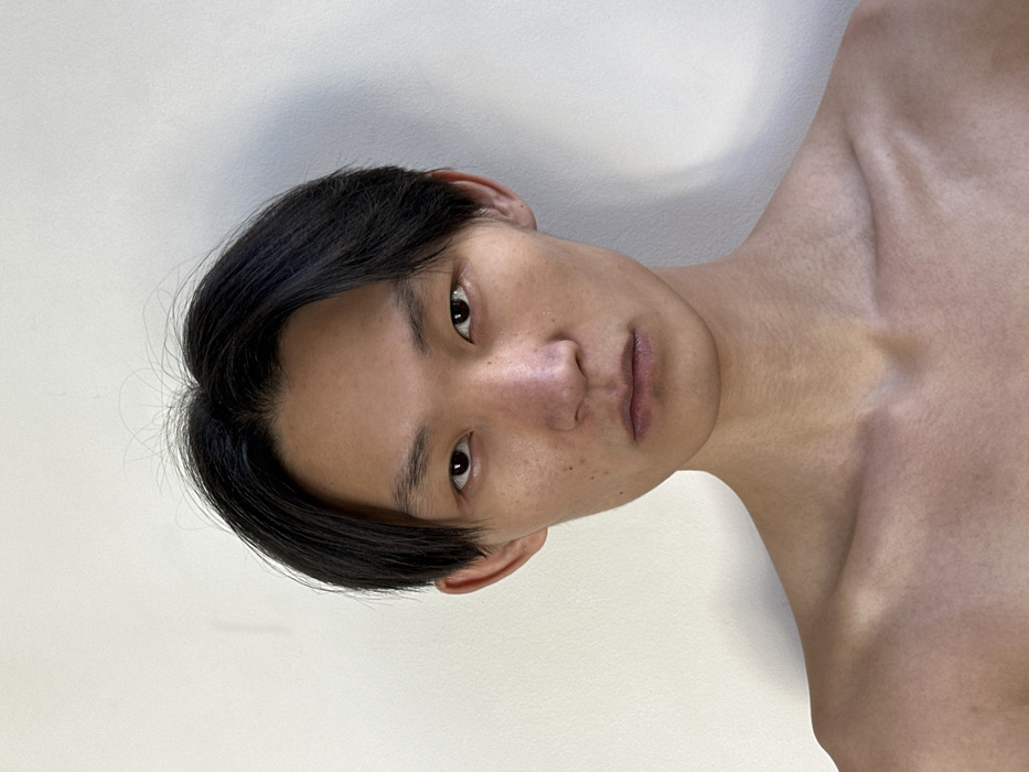 A head-and-shoulders portrait depicts an individual identified as an East Asian male, captured from the mid-chest upward, against a plain, light-colored wall. The subject's dark hair is parted in the middle and styled to frame the face. His dark eyes are open, directed forward, and hold a neutral expression. The mouth is closed in a relaxed, straight line. The individual is shirtless, revealing bare shoulders, prominent collarbones, and upper chest. The skin is a light to medium tone, exhibiting minimal blemishes. Even, frontal lighting illuminates the subject, casting subtle shadows. The scene is situated indoors, within an unspecified location in Paris, France.