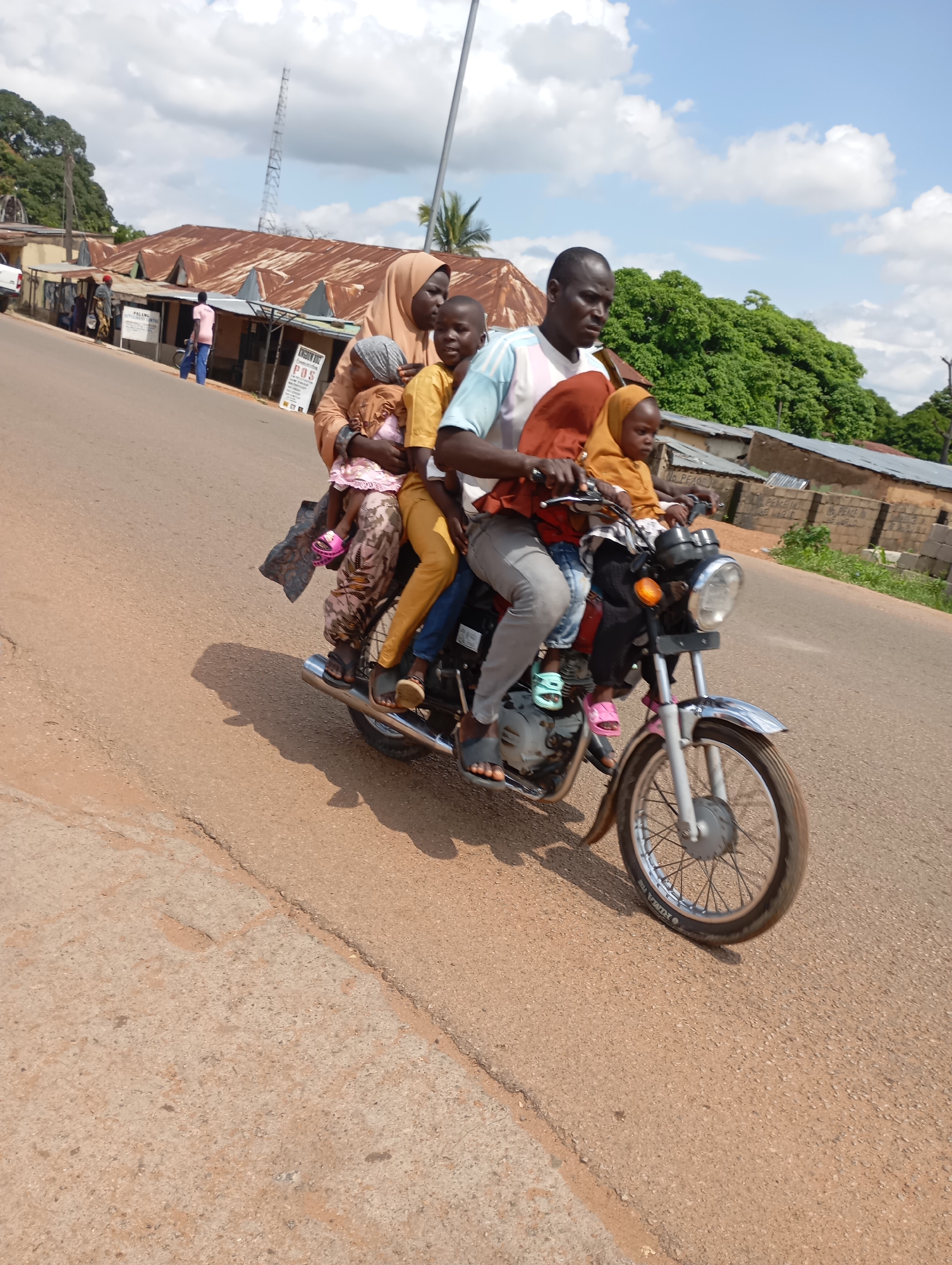 In Shendam, Nigeria, a group of five people, including at least two adults and three children, are riding on a motorcycle. A man is driving, with a woman behind him holding a baby. Two other children are also on the motorcycle, one positioned in front of the driver and another between the driver and the woman. The motorcycle is traveling on a paved road lined with buildings on the right side of the image. The buildings appear to be single-story structures with corrugated metal roofs. Trees and vegetation are visible in the background, and the sky is partly cloudy. The scene captures a common mode of transportation in the region.