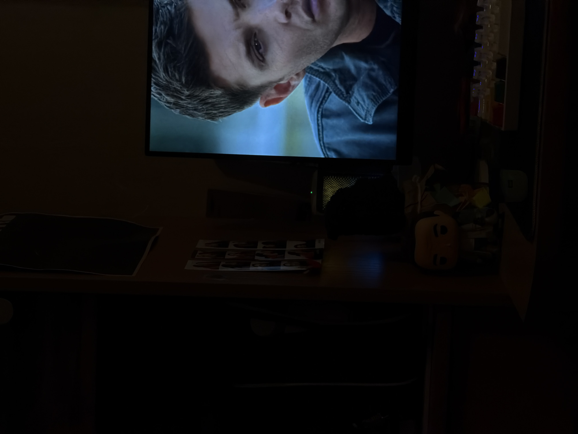The image shows a desk with a monitor displaying a close-up of a man's face and shoulder. To the right of the monitor is a keyboard. On the desk are also a black rectangular object, a sheet of images, and a small doll. The surroundings are dark. There is no indication of the location being Gamovo, Russia.