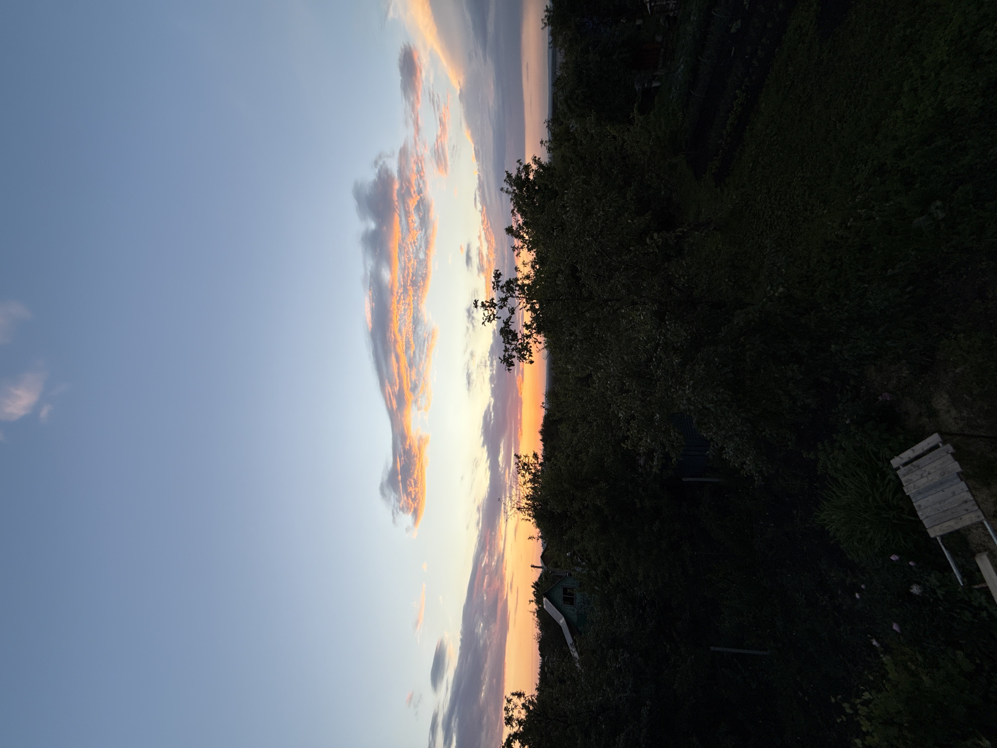 A wide shot captures a rural landscape at dusk. The sky is a gradient of pale blue at the top, transitioning to hues of orange and pink near the horizon where the sun has recently set. Wispy clouds are illuminated by the last rays of sunlight, creating streaks of color across the sky. In the foreground, dense green trees and foliage dominate the scene, casting dark shadows. Peeking through the trees is a small, dark green house with a pitched roof. The ground below is covered in vegetation, with some darker, unidentifiable areas that appear to be cultivated plots or dense undergrowth. A wooden structure, possibly a ramp or stairs, is visible in the lower left quadrant of the image. The overall atmosphere is serene and tranquil, indicative of a quiet evening in the countryside.