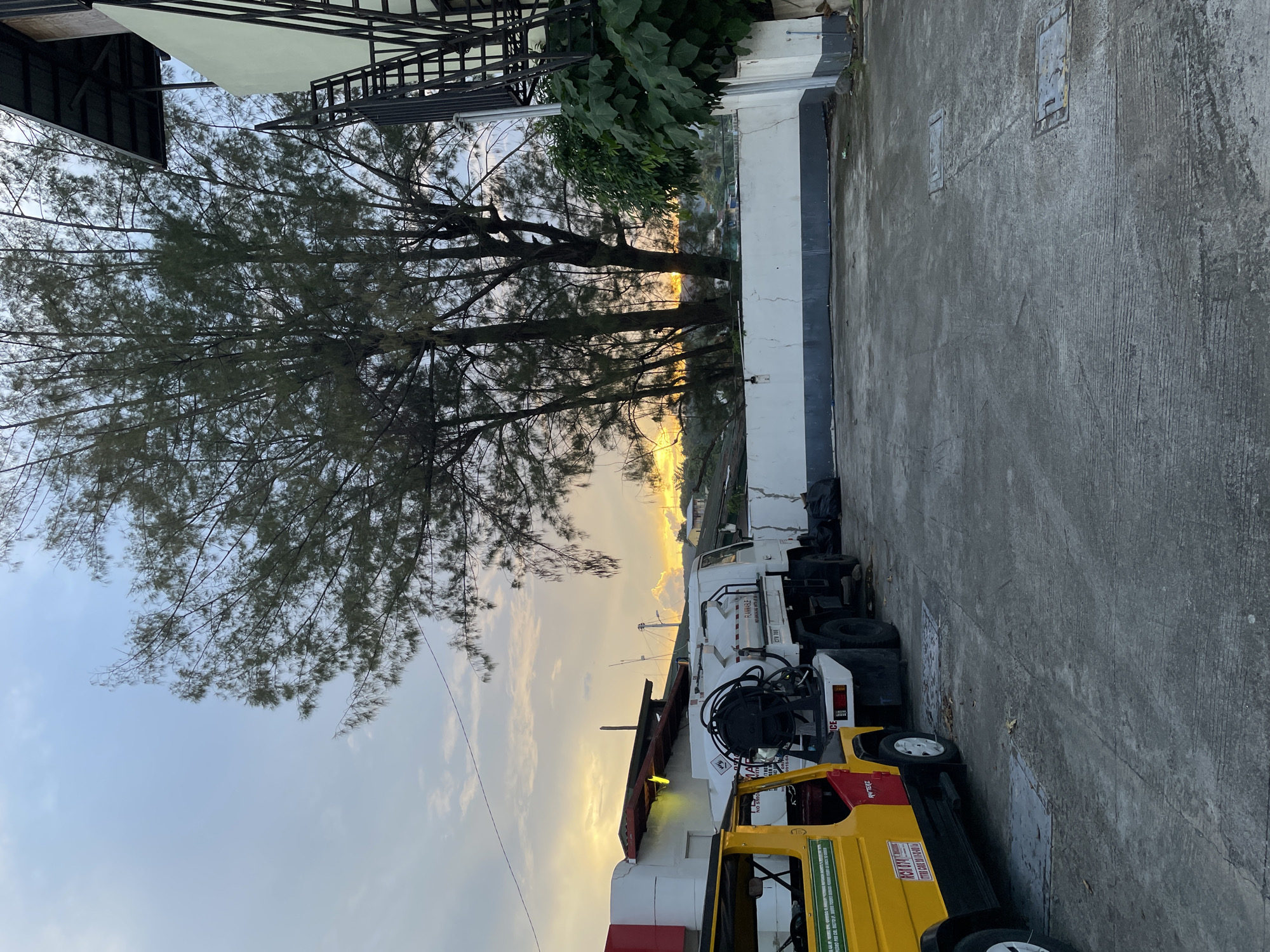 The image depicts an outdoor scene in Davao City, Philippines, likely during either sunrise or sunset, as indicated by the warm glow in the sky. In the foreground, a concrete pavement area is visible, with some metal manhole covers. Two vehicles are parked on the pavement: a yellow and red jeepney on the left, and a white truck with a large tank on the right. The jeepney has visible markings and a license plate that appears to read "RGN 074". The white truck has markings indicating it is a "FLAMMABLE" vehicle and has a hose reel attached to its tank. Behind the vehicles, a low white wall with a dark gray trim runs horizontally across the image. Beyond the wall, a dense cluster of trees with thin branches and needles dominates the mid-ground. In the background, a building with a light-colored facade and an external metal staircase is partially visible on the right. The overall impression is that of an industrial or commercial area.