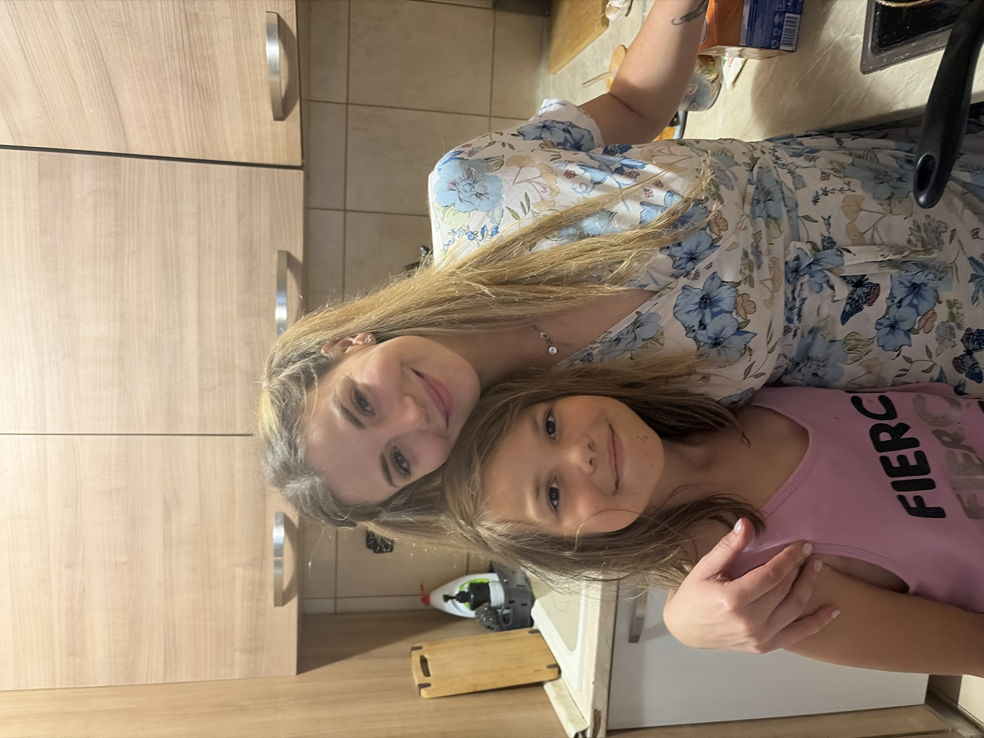 This is a waist-up, eye-level, indoor, medium shot of two females in a kitchen. The female in the foreground, a young girl with brown hair, is facing the camera and smiling. She is wearing a pink tank top with the word "FIERCE" in black lettering. The female behind her, a young woman with blonde hair styled in pigtails, is also facing the camera and smiling. She is wearing a floral print wrap dress with blue and white flowers. Her arm is around the girl's shoulders. The kitchen features light wood cabinets and countertops. A sink, a cutting board, and a bottle of dish soap are visible on the counter.