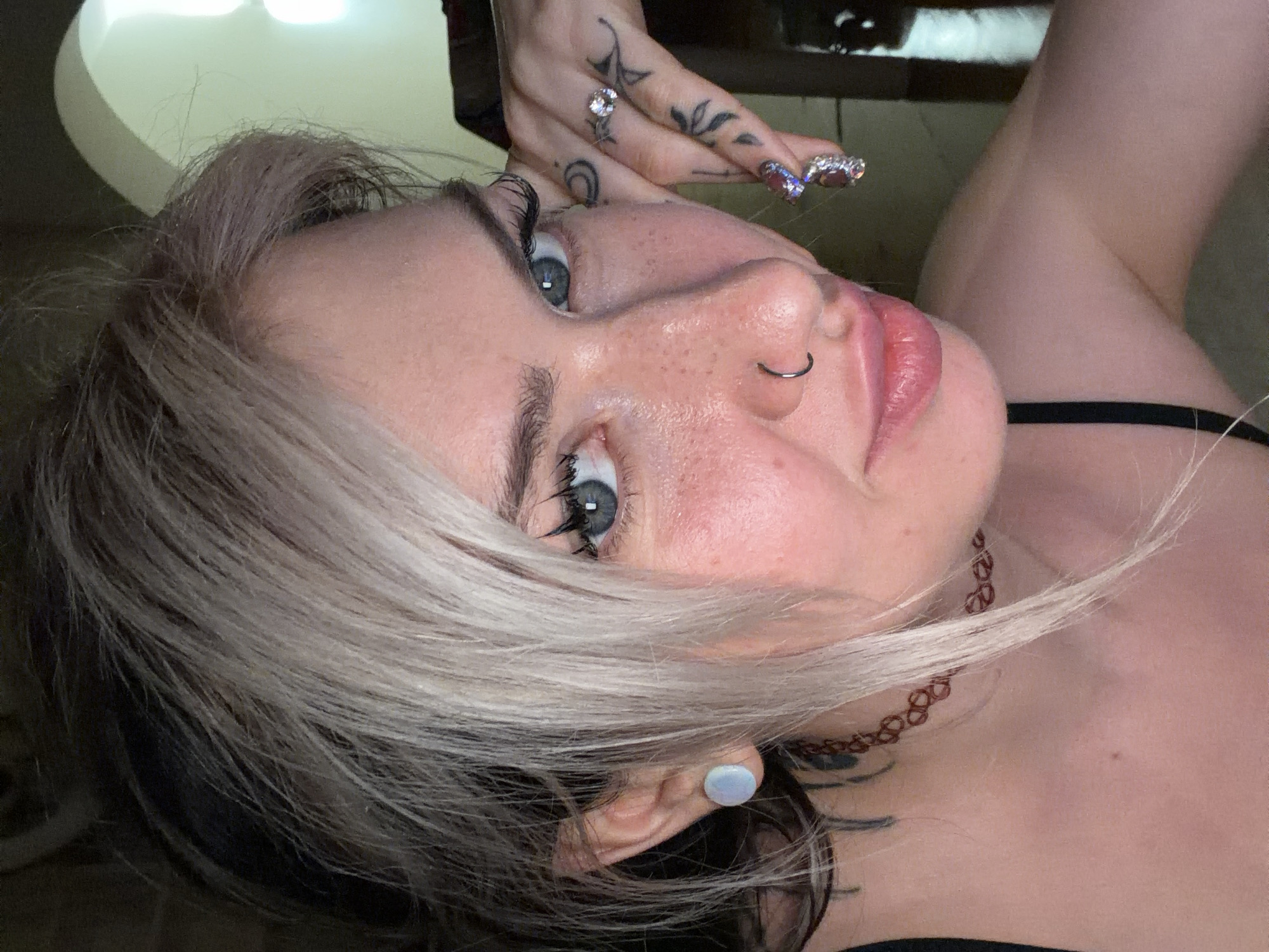A close-up, angled portrait of a person with fair skin, freckles, and light grey/blonde hair with dark roots. They have blue eyes, a nose ring, and their hand is raised near their forehead, displaying rings and tattoos on their fingers. They are wearing a thin, black strap and a brown choker. The background is out of focus, with a hint of a light fixture and an obscured dark area. The location in the city of Budva, Montenegro, cannot be determined from the image.