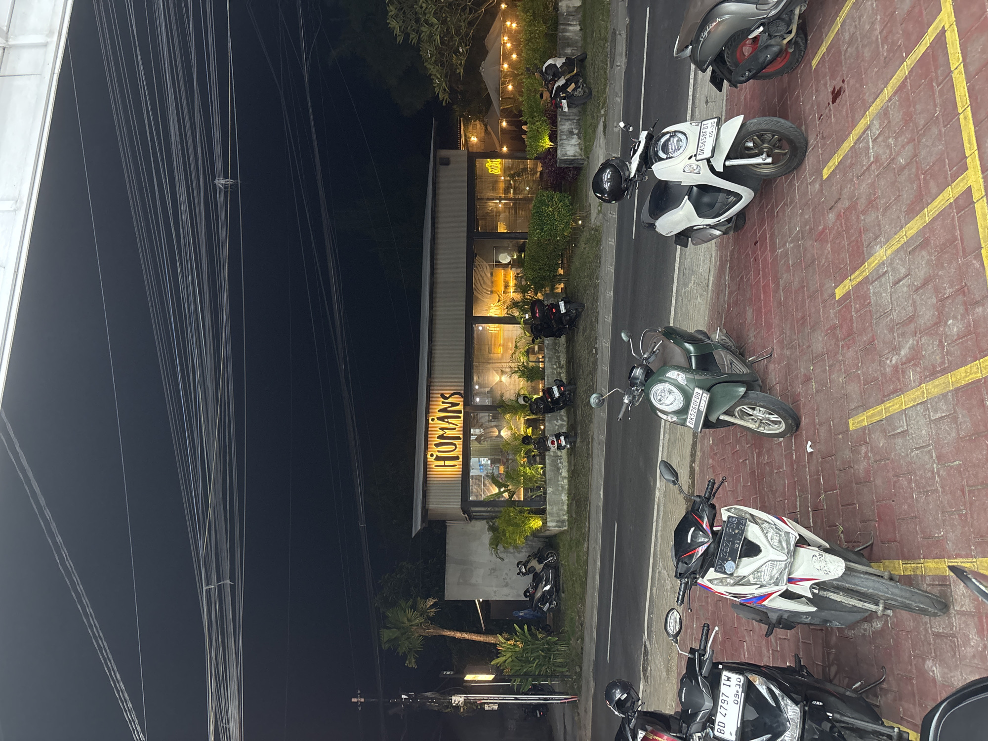 Here is a description of the image:
The image shows a nighttime view of "Humans" restaurant in Ungasan, Indonesia. The restaurant has a large, illuminated sign. Several motorbikes are parked along the side of the road and on the sidewalk in front of the restaurant. The restaurant's interior is lit and visible through the large glass windows. Overhead power lines run across the top left of the image. The sidewalk is made of red pavers with yellow parking lines. The street is asphalt and appears wet. Trees and shrubs are also present around the building. The scene is lit by streetlights and the restaurant's lighting.