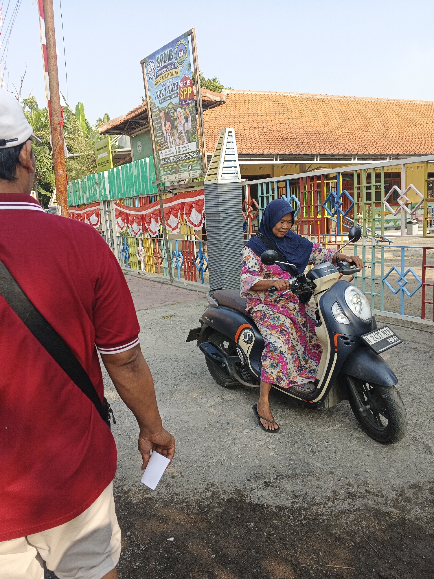 The image depicts a street scene in Krian, Indonesia, focusing on a woman riding a motorcycle. She is wearing a blue hijab and a colorful floral dress, and appears to be smiling. The motorcycle is dark-colored with some beige accents and has a license plate that reads "L 2437 WBK". In the foreground, a person wearing a red polo shirt and a white cap is shown from the back, holding a small white piece of paper. Behind the woman on the motorcycle, there's a building with an orange tiled roof, a colorful fence, and a billboard advertising "SPMB" for the academic year 2027-2028. The environment appears to be a sunny day in a suburban or semi-urban area, possibly near a school or educational institution given the presence of the billboard and colorful fencing.